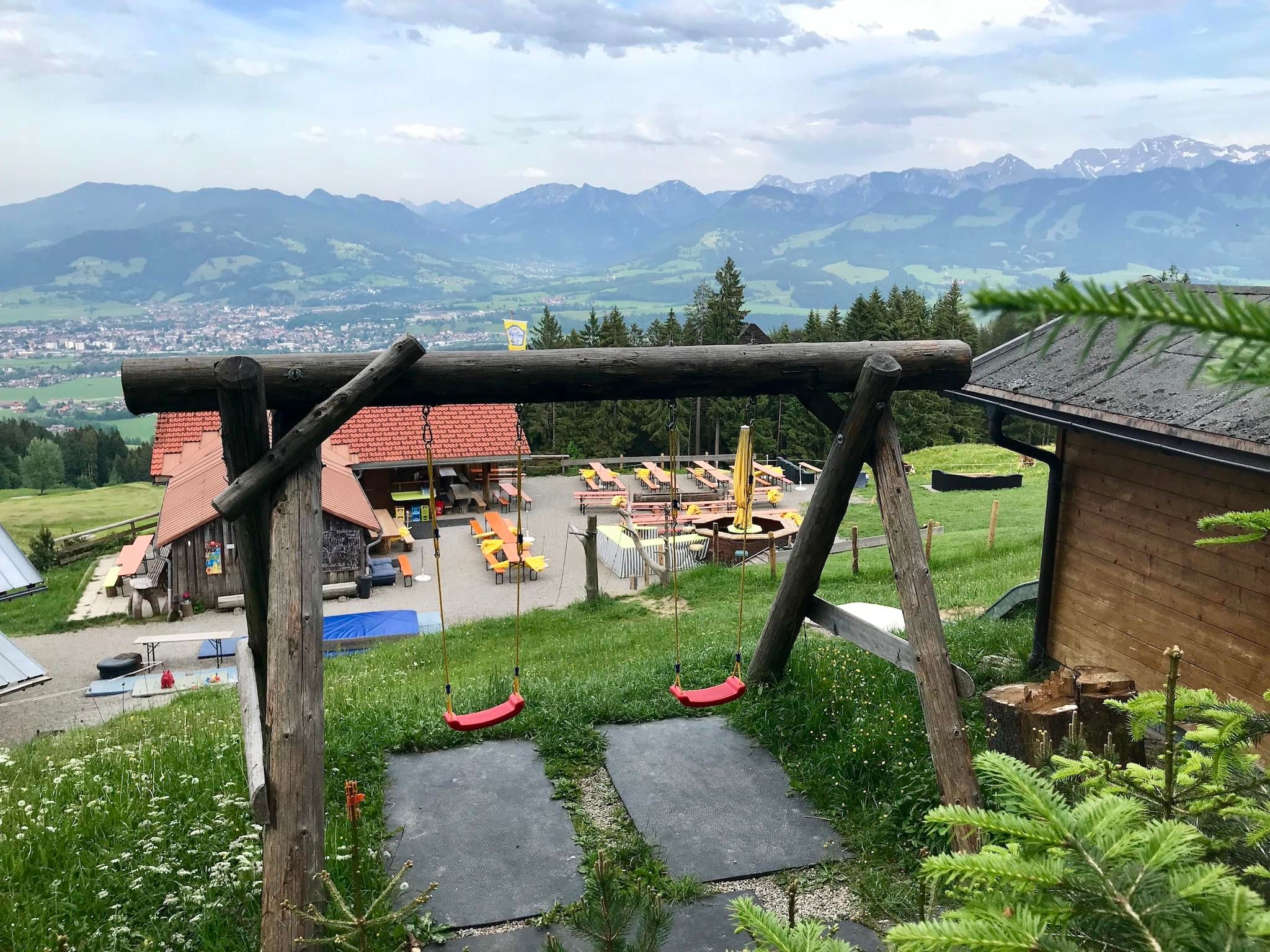 Eine Holzschaukel vor einer Alpe mit Sitzbänken eröffnet einen beeindruckenden Blick ins Tal und auf die umliegende Bergwelt. Die Szene wirkt gemütlich, einladend und familienfreundlich.