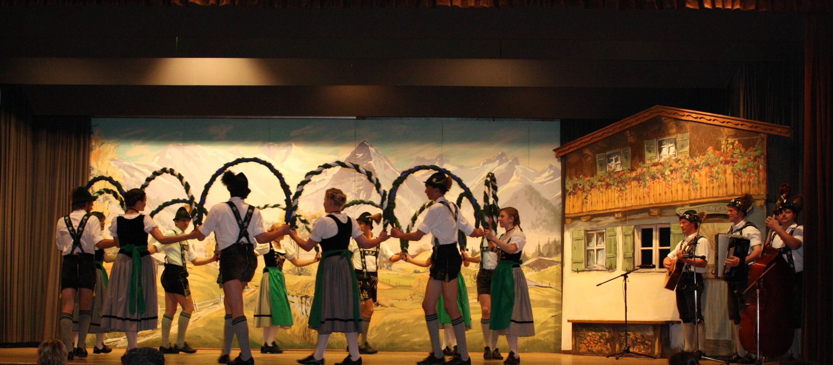 Bühnenaufführung mit Trachten-Tanzgruppe, Musikern und gemalter Bergkulisse.