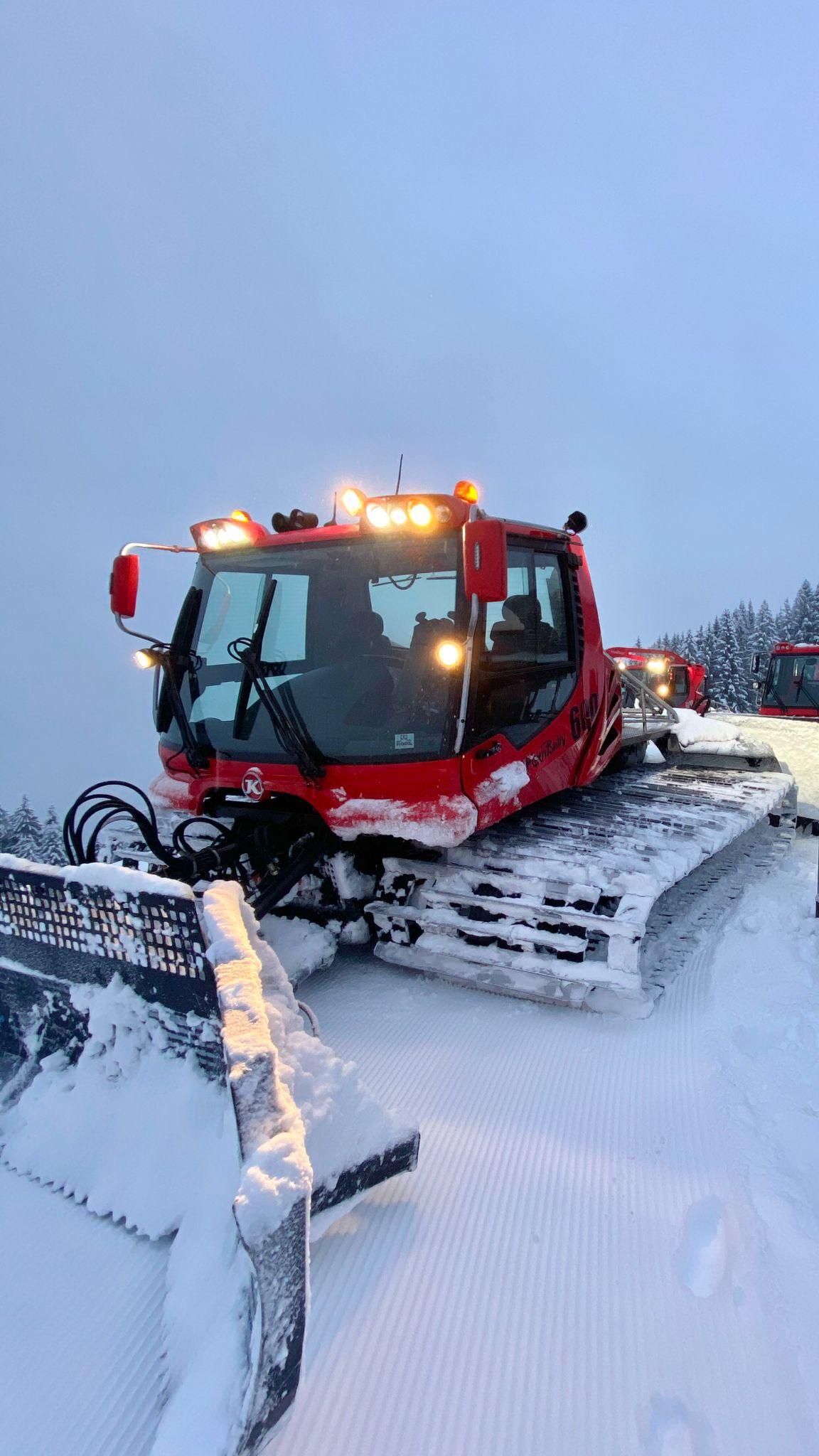Eine rote Pistenraupe steht auf einer verschneiten Piste im Gebirge. Ihre Scheinwerfer und Warnlichter leuchten.