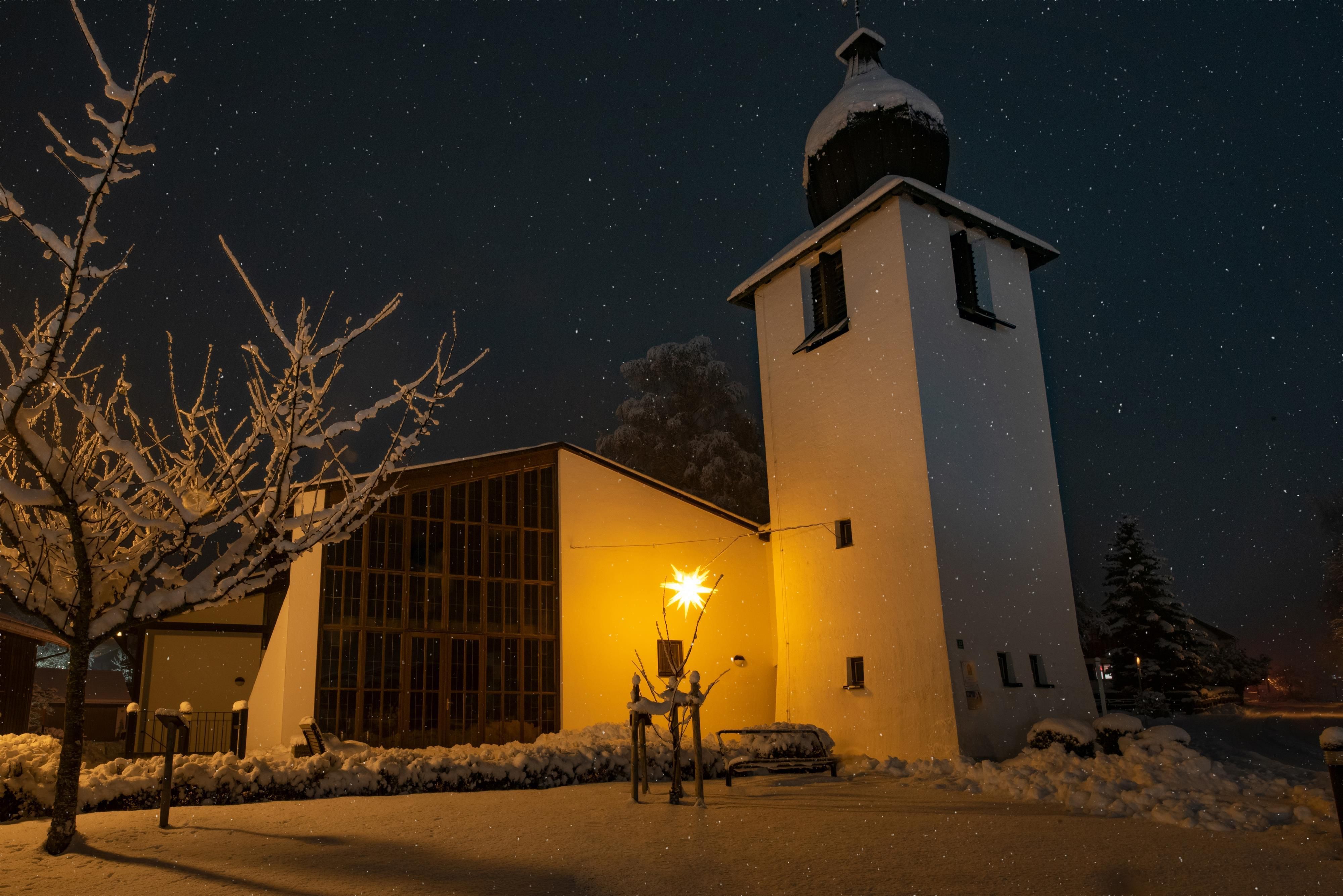Eine schneebedeckte Kirche mit einem Uhrturm im Dunklen. Der Himmel ist klar und es fallen kleine Schneeflocken.