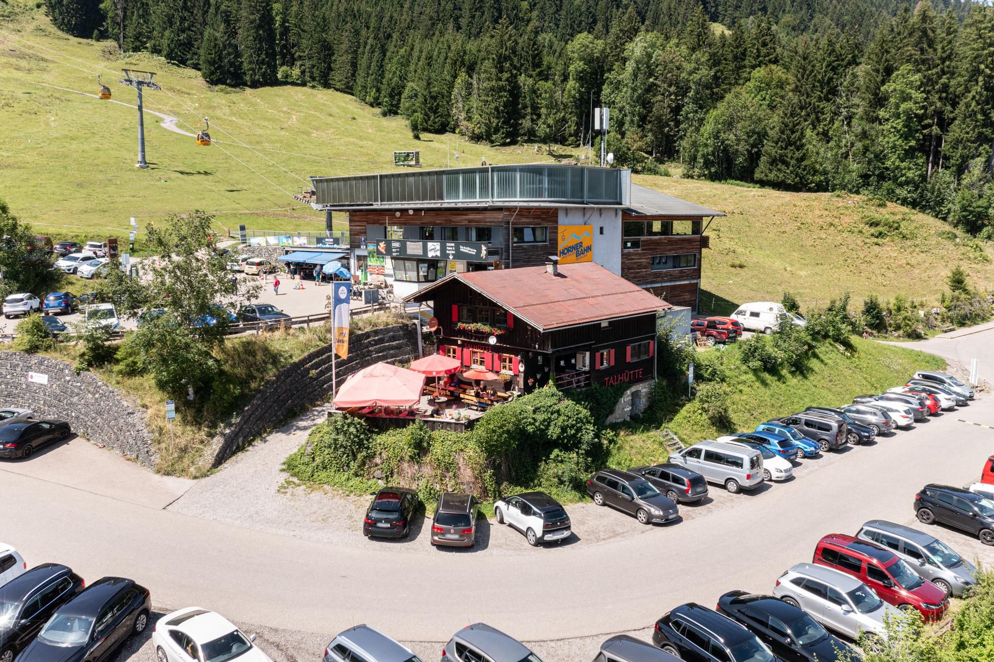 Übersicht der Talhütte in Bolsterlang an einem sonnigen Tag. Parkplatz mit vielen Autos. Gebäude mit rotem Dach und Außenbereich mit Sonnenschirmen.