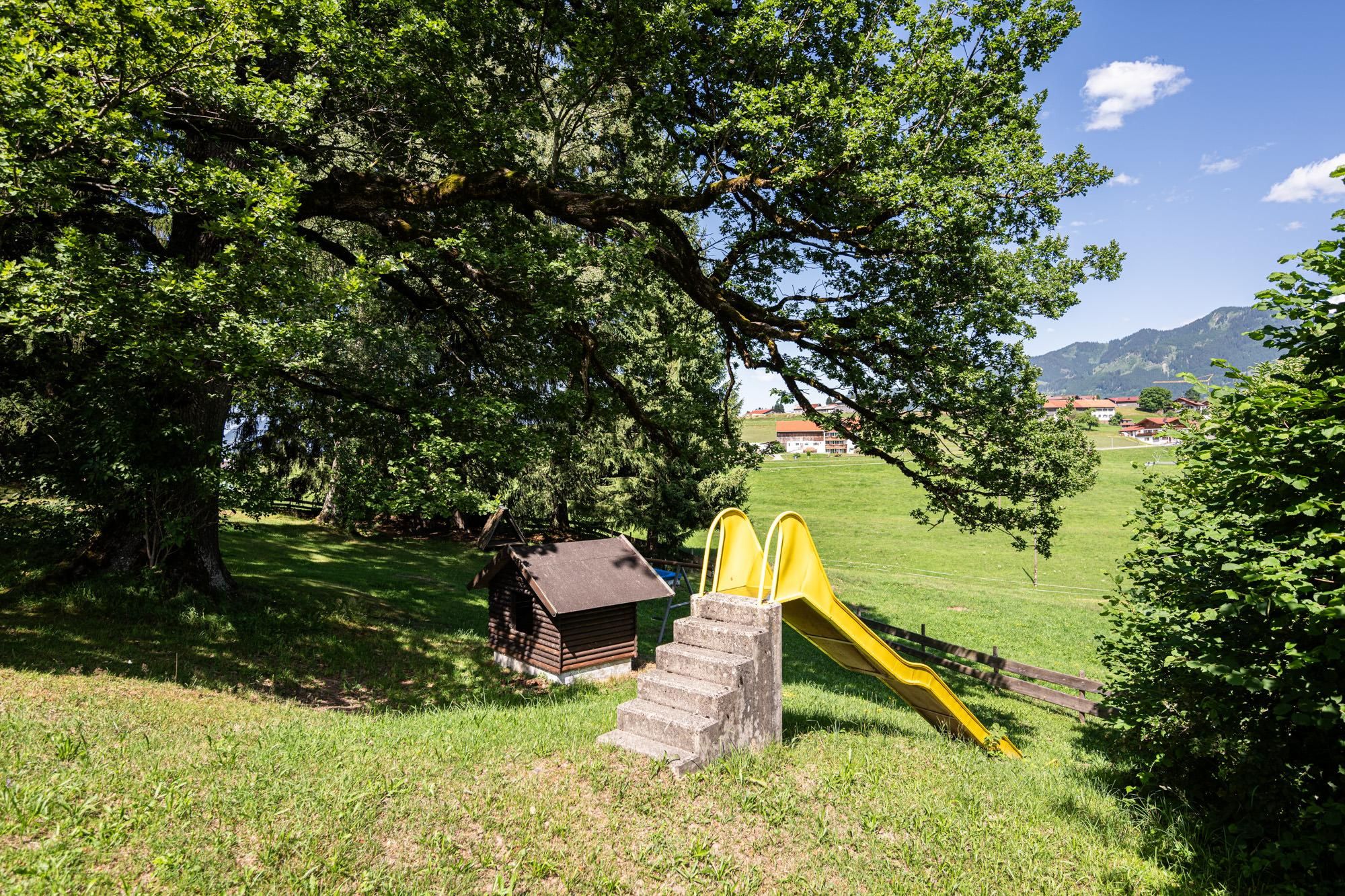 Spielplatz Spöckwiese. Gelbe Rutsche mit Steinstufen. Kleines Holzhaus unter einem großen Baum. Grüne Wiese, Holzzaun und Häuser im Hintergrund.