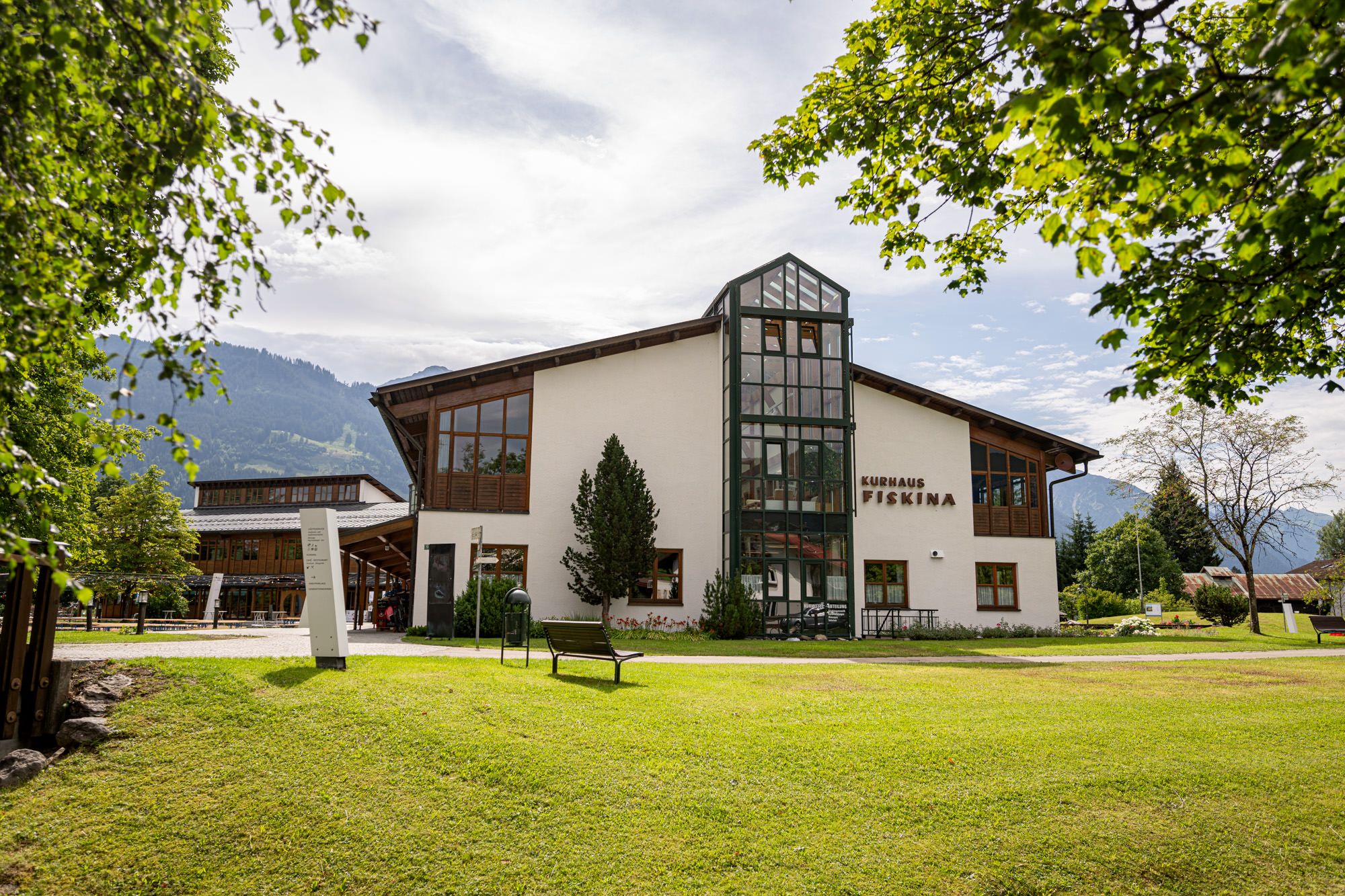 Zweigeschossiges, modernes Kurhaus in Fischen. Grüne Gärten umgeben das Gebäude, dahinter die Berge.