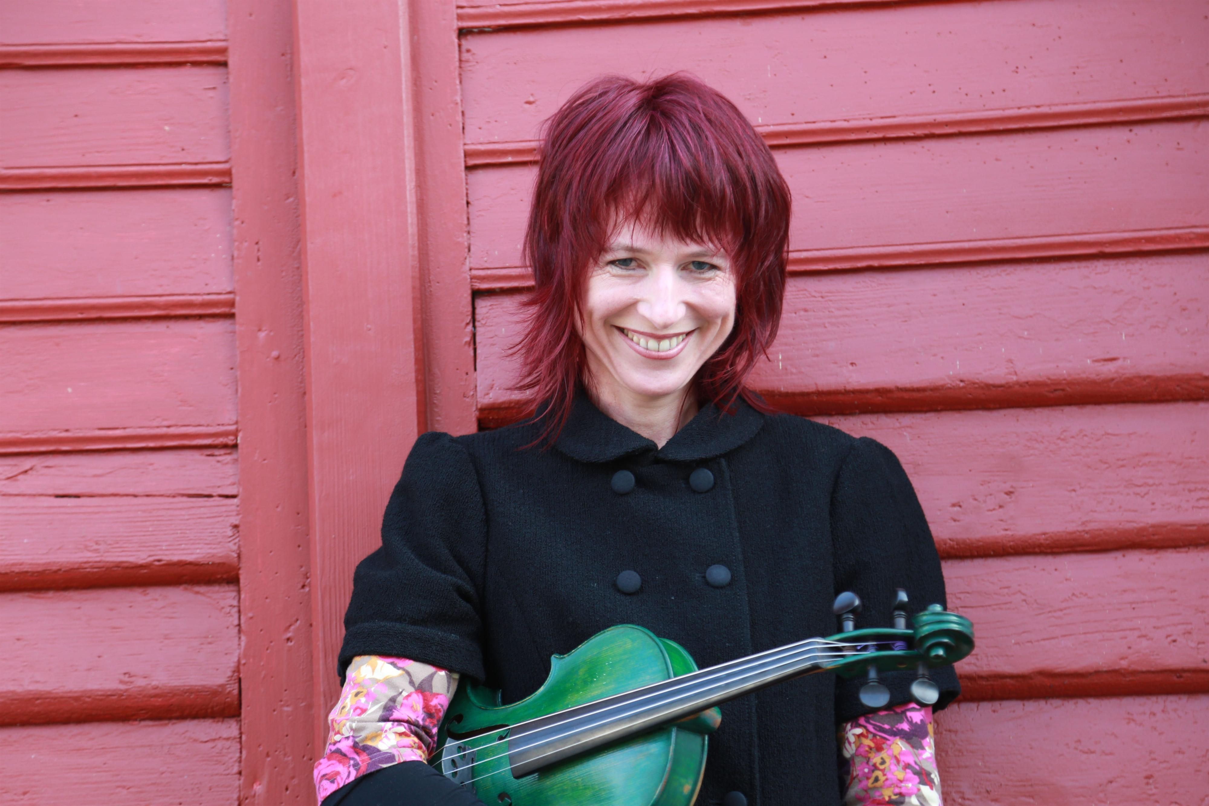Eine Frau mit leuchtend roten Haaren sitzt vor einer rot gestrichenen Holzwand und lächelt warm in die Kamera. In ihren Armen hält sie eine grüne Geige