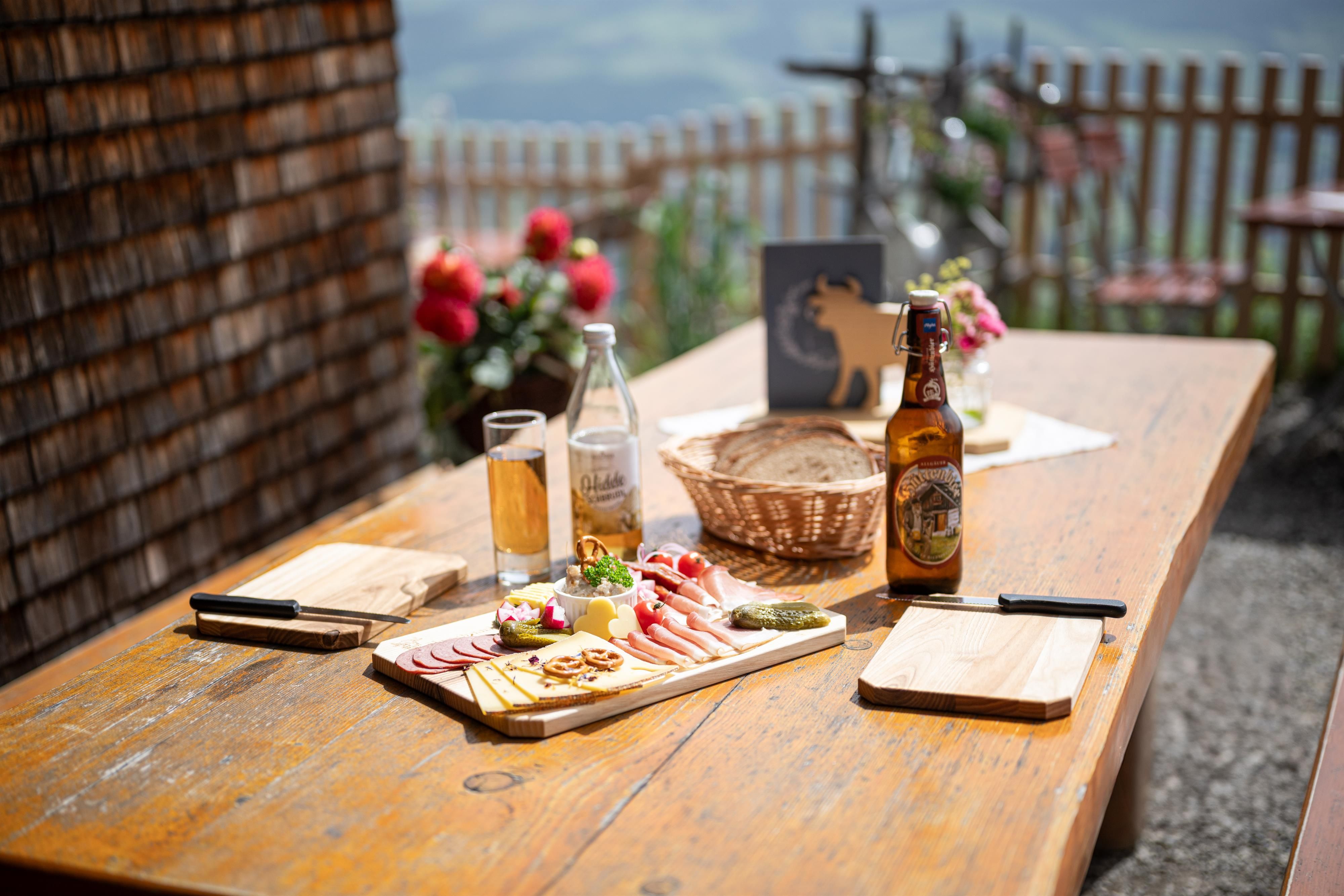 Auf einem Holztisch steht eine Brotzeitplatte bestehend aus Wurst, Käse und Dekoration. Dahinter befindet sich ein Brotkorb. Auch zwei Brotzeitbretter und Getränke stehen auf dem Tisch. Im Hintergrund der Blick in ein grünes Tal.