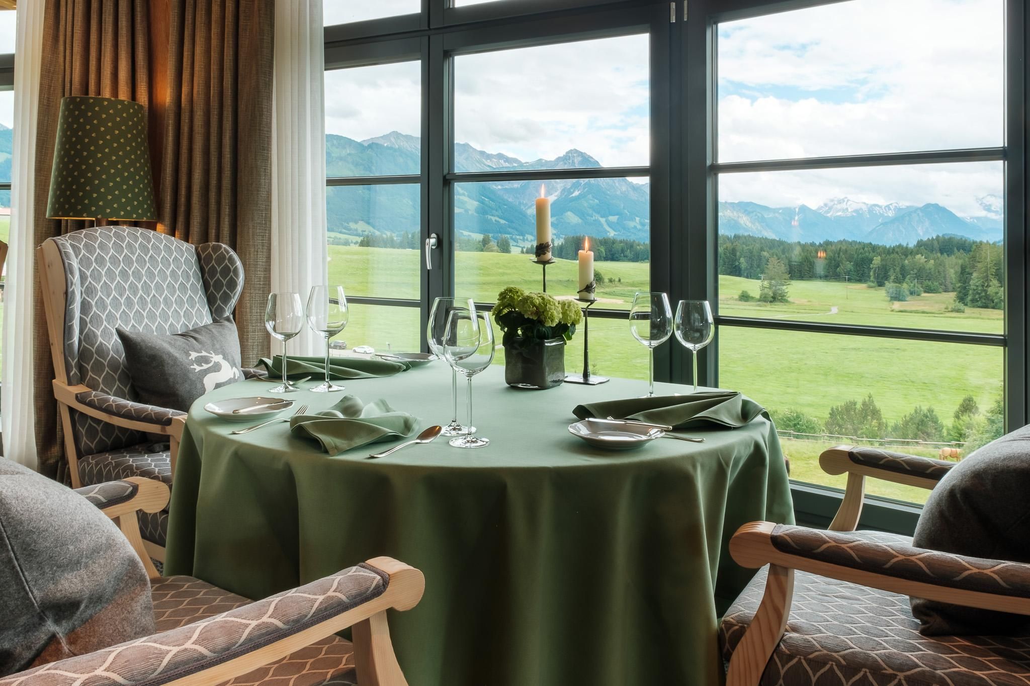 Ein elegantes Esszimmer mit Blick auf grüne Wiesen und Berge. Der Tisch ist schön gedeckt und es gibt Kerzen und Gläser.