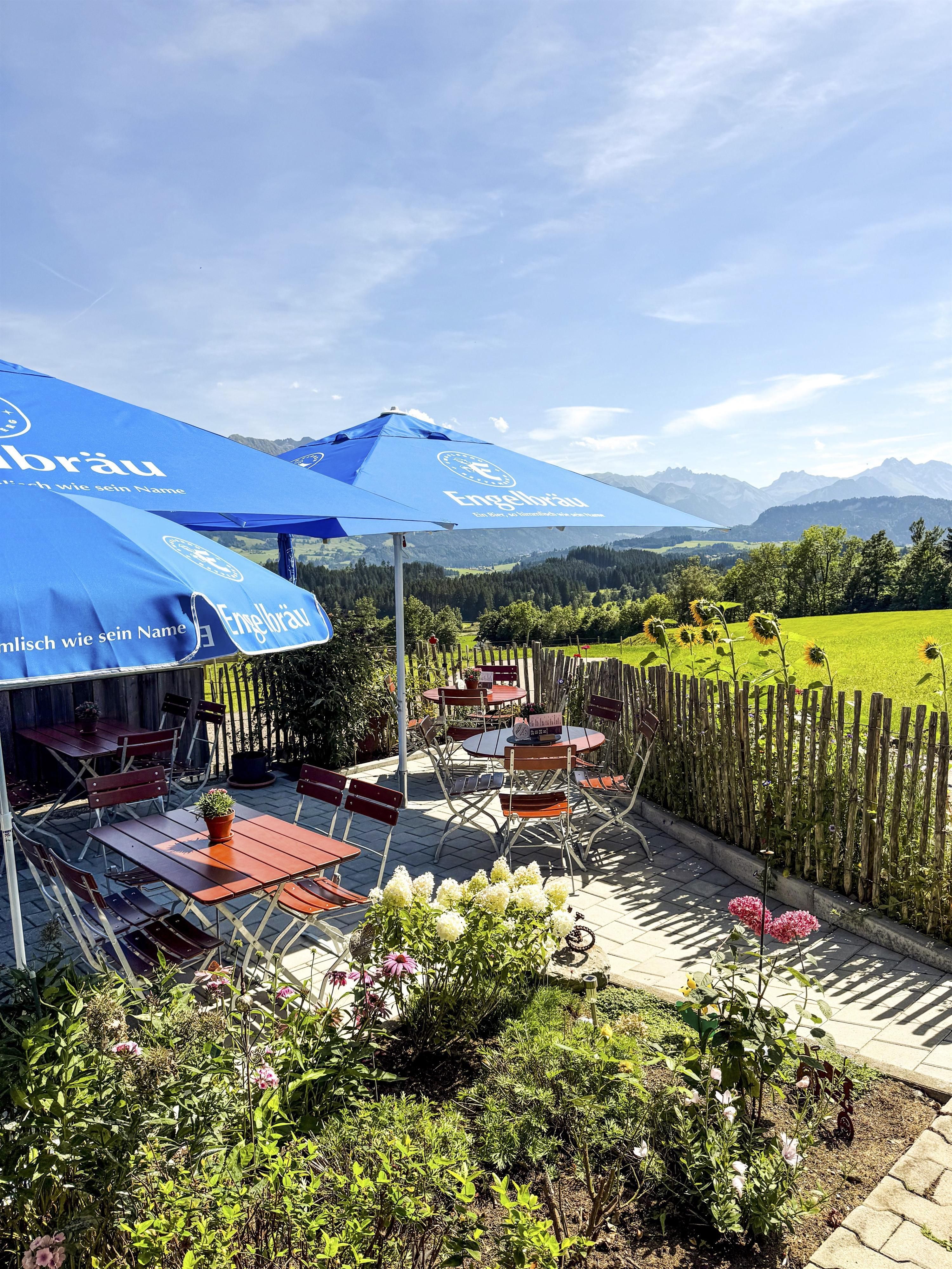Ein gemütlicher Außenbereich eines Restaurants mit Holzstühlen und Tischen. Im Hintergrund sind grüne Wiesen und Berge unter einem klaren blauen Himmel zu sehen.