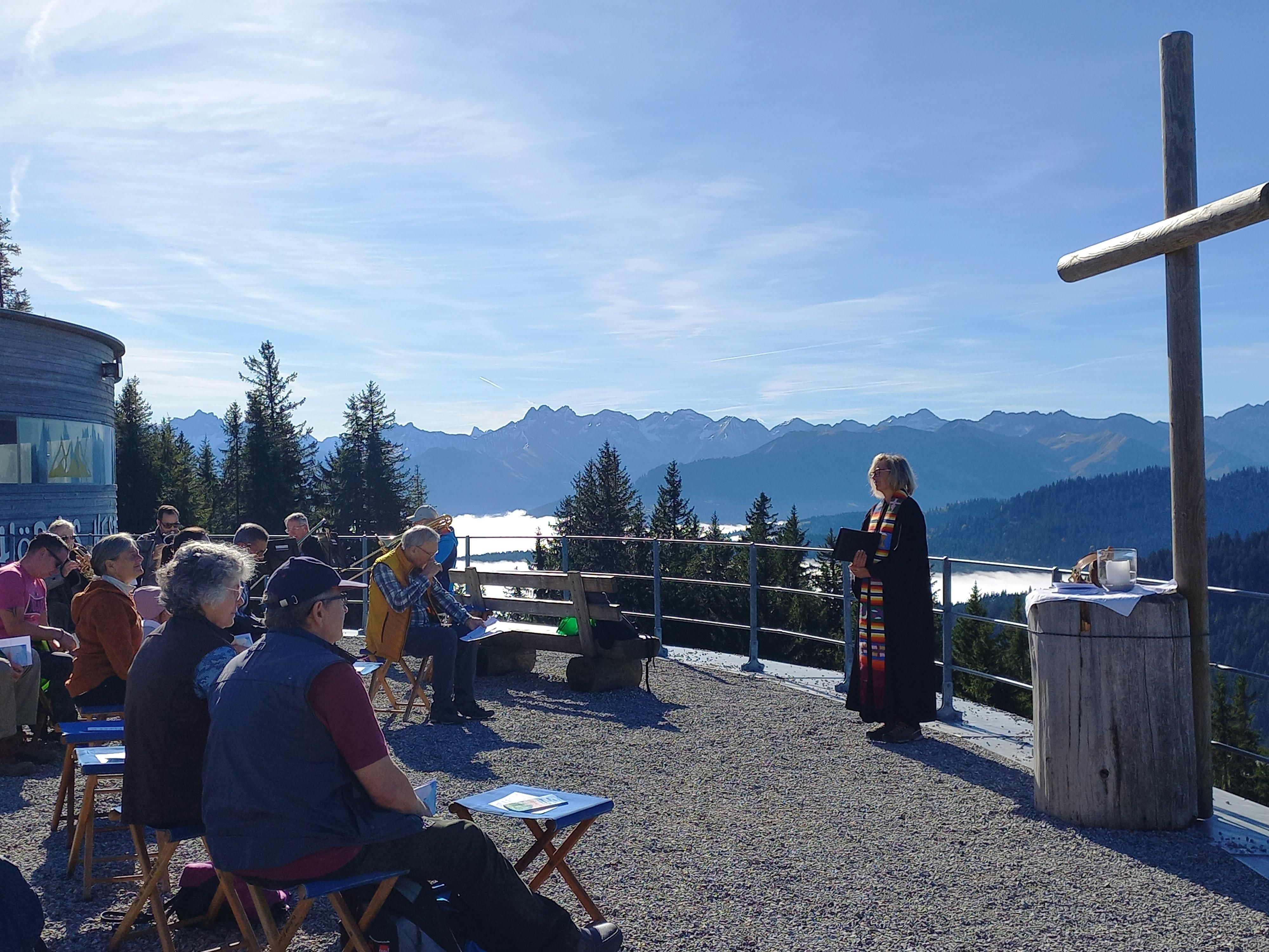 Eine Person steht vor einer Gruppe von Menschen auf einem Aussichtspunkt in den Bergen. Im Hintergrund sind hohe Berge und ein blauer Himmel zu sehen.