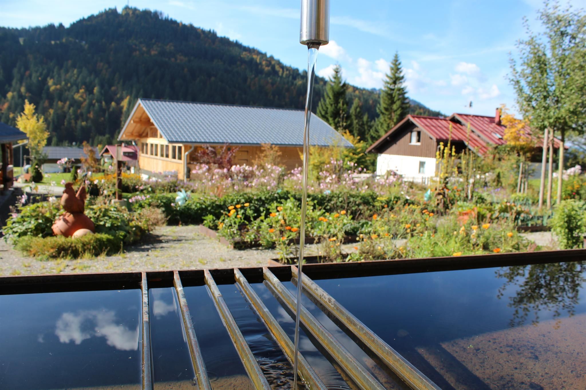 Wasserstrahl aus Metallrohr fällt in dunkles Wasserbecken, bunte Blumenbeete mit Häusern im Hintergrund.