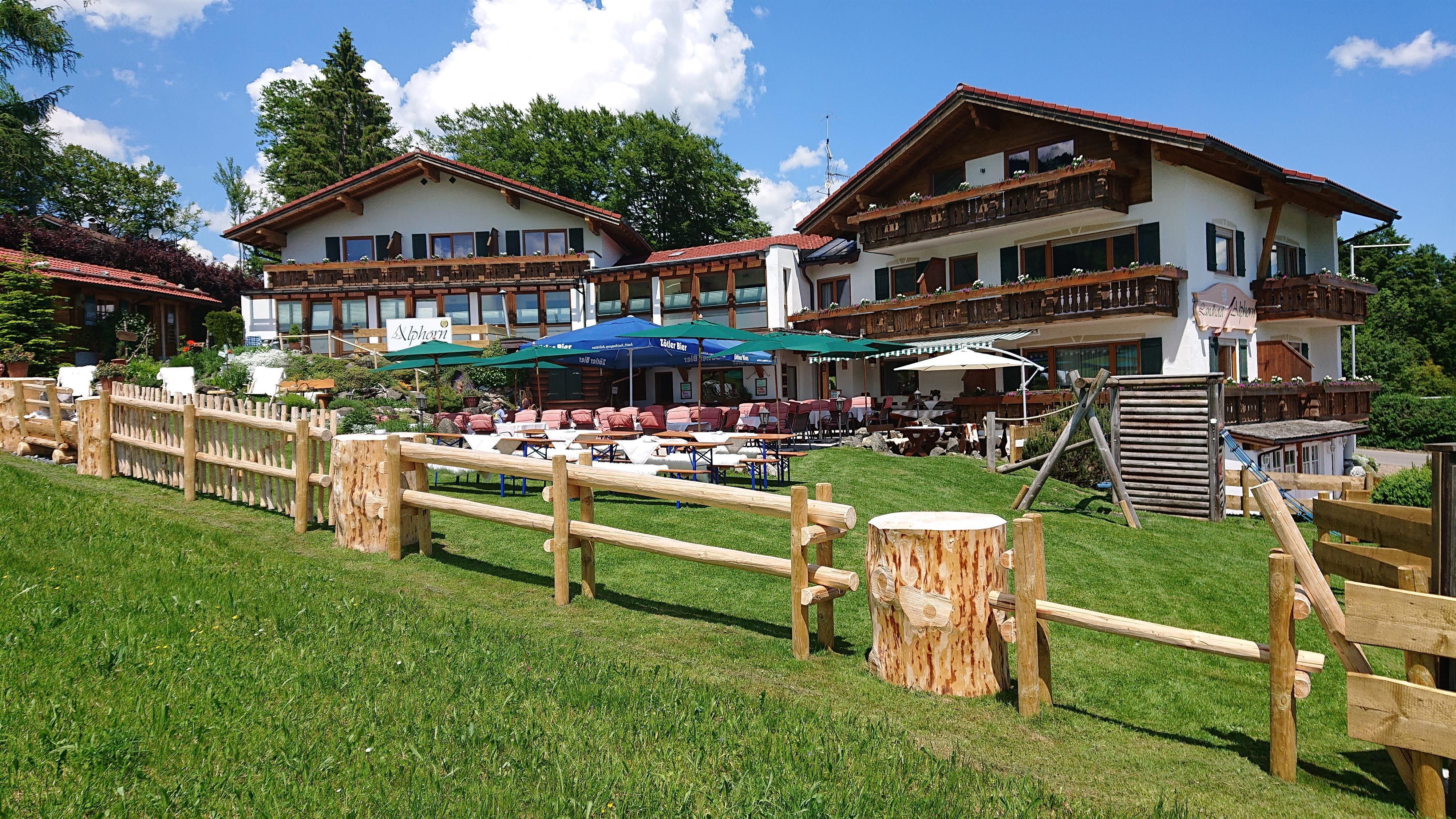 Zwei traditionelle bayerische Gebäude mit Holzbalkonen und roten Ziegeldächern stehen auf einem grünen Hang.