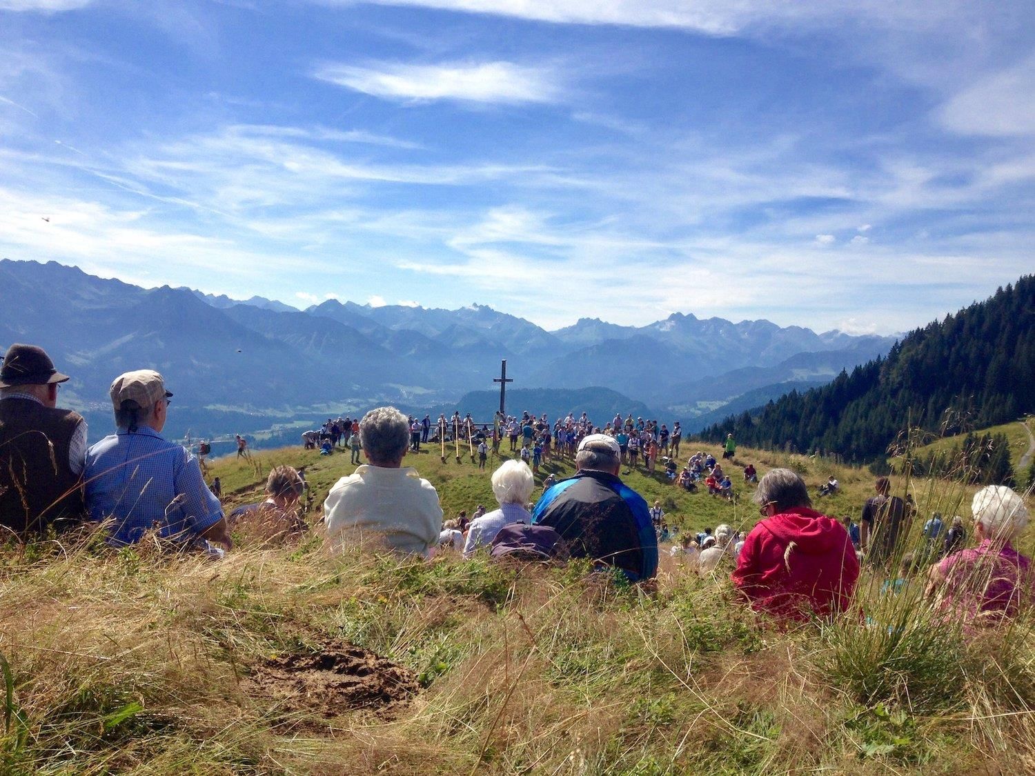 Menschen sitzen auf einer grünen Wiese und blicken auf ein Holzkreuz. Dahinter erstreckt sich eine weite Landschaft mit bewaldeten Hügeln und Bergen.
