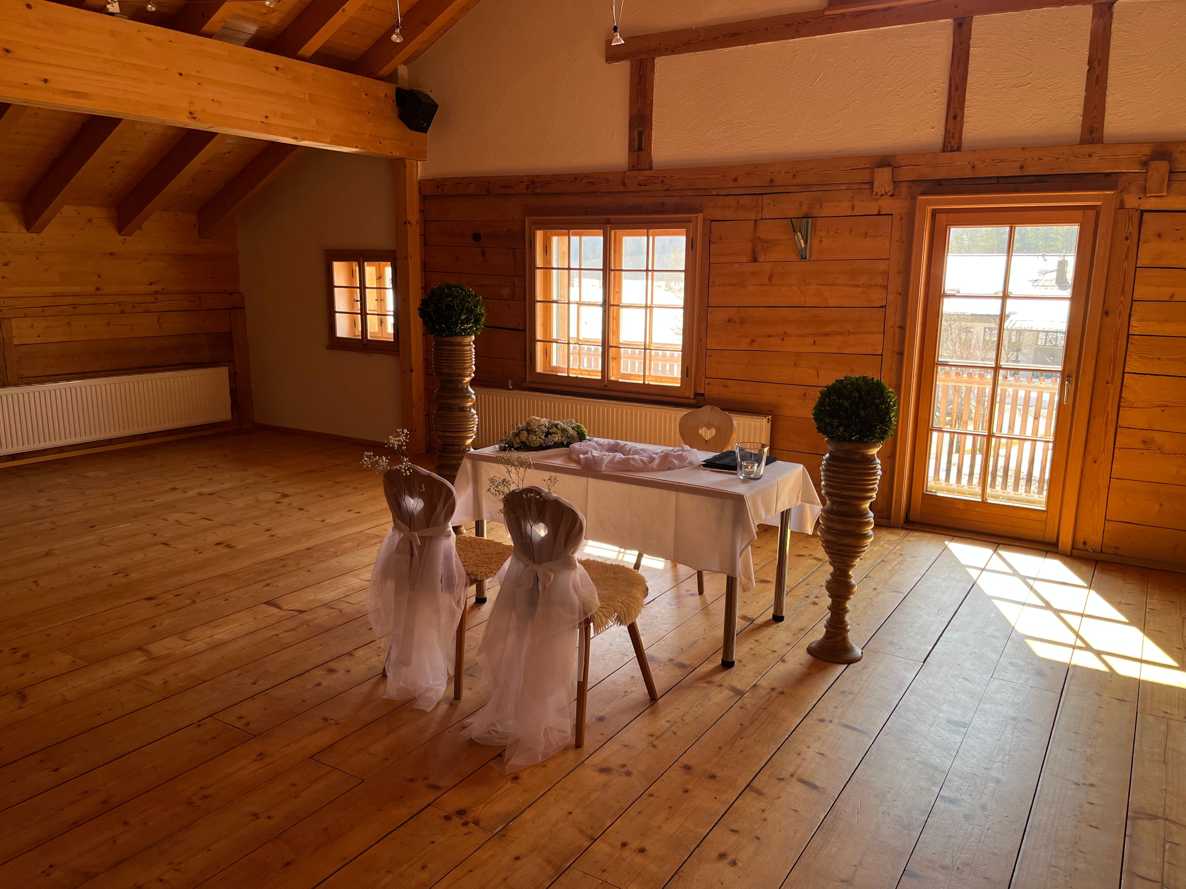 Großer Raum "Weitblick" mit offenem Dachstuhl und viel Holz. Festlich dekorierter Trautisch vor hellen Fenstern.