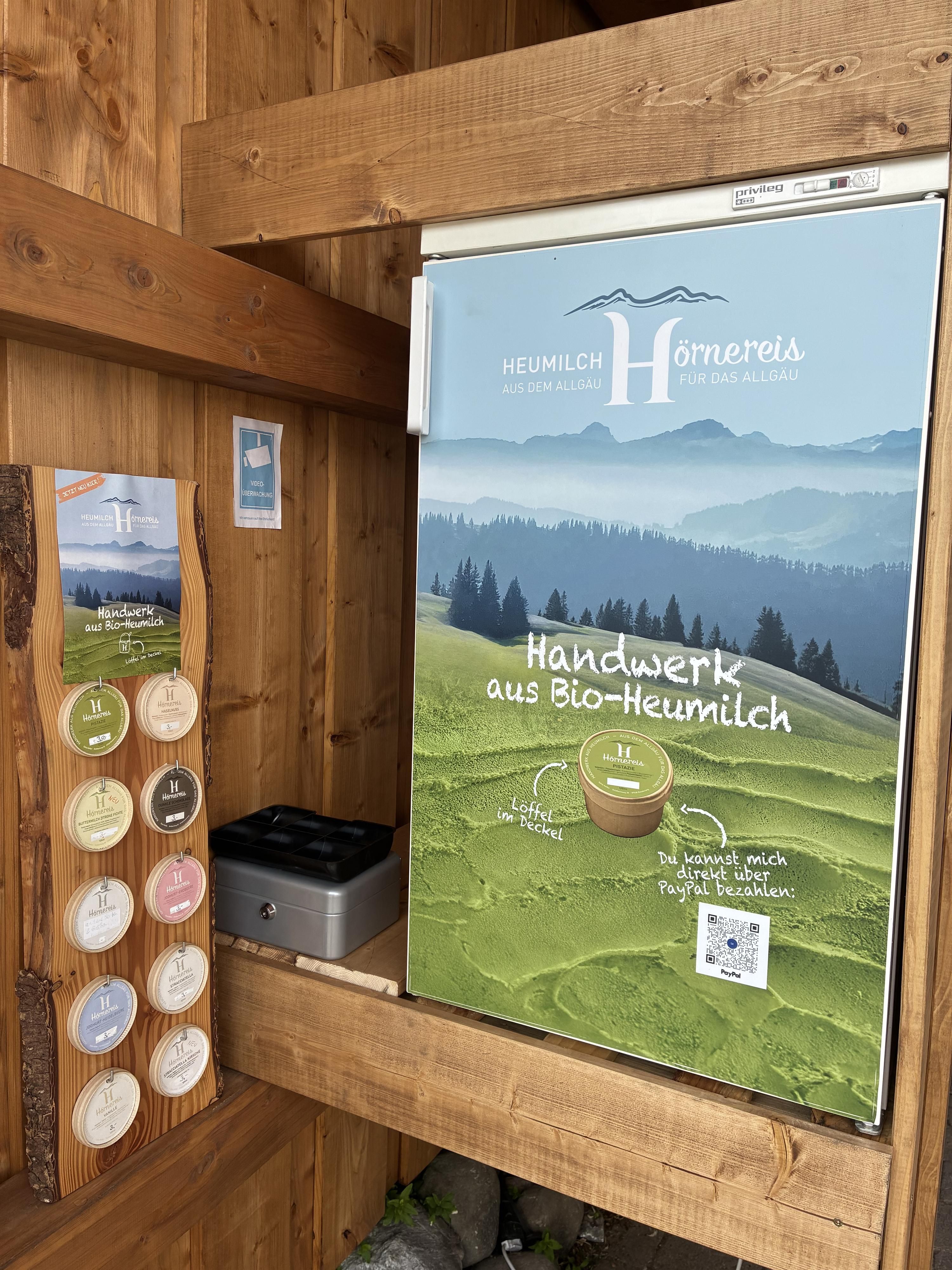 Ein Kühlschrank aus Holz mit einem Werbeschild für handwerkliche Produkte aus Bio-Heumilch steht an einer Wand. Daneben befindet sich eine Übersicht mit verschiedenen angebotenen Produkten.