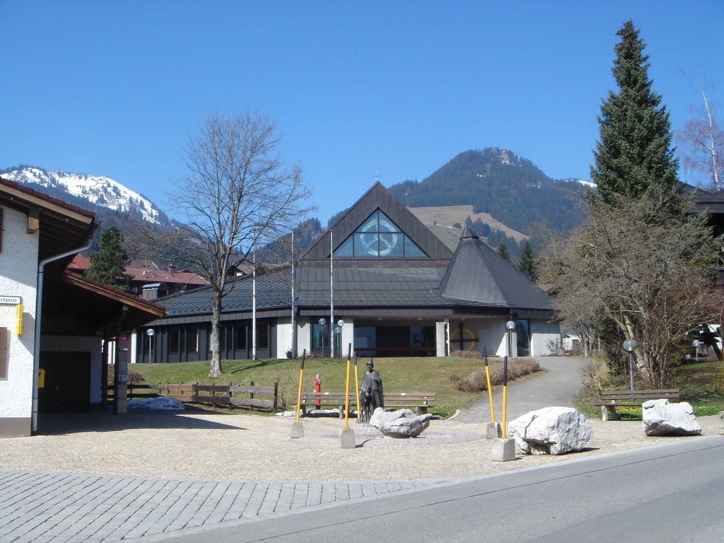 Moderne Kirche in Obermaiselstein mit spitzem Dach vor Bergkulisse, Bänke und Statue im Vordergrund.
