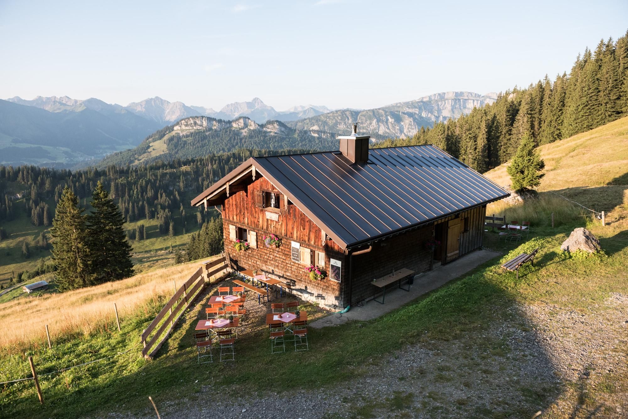 <p>Bewirtschaftete Hütte  mit dunklem Dach, Tische und Stühle draußen, Blumenkästen an den Fenstern, umgeben von gelblichen Hängen, Bäumen und Bergen.</p>