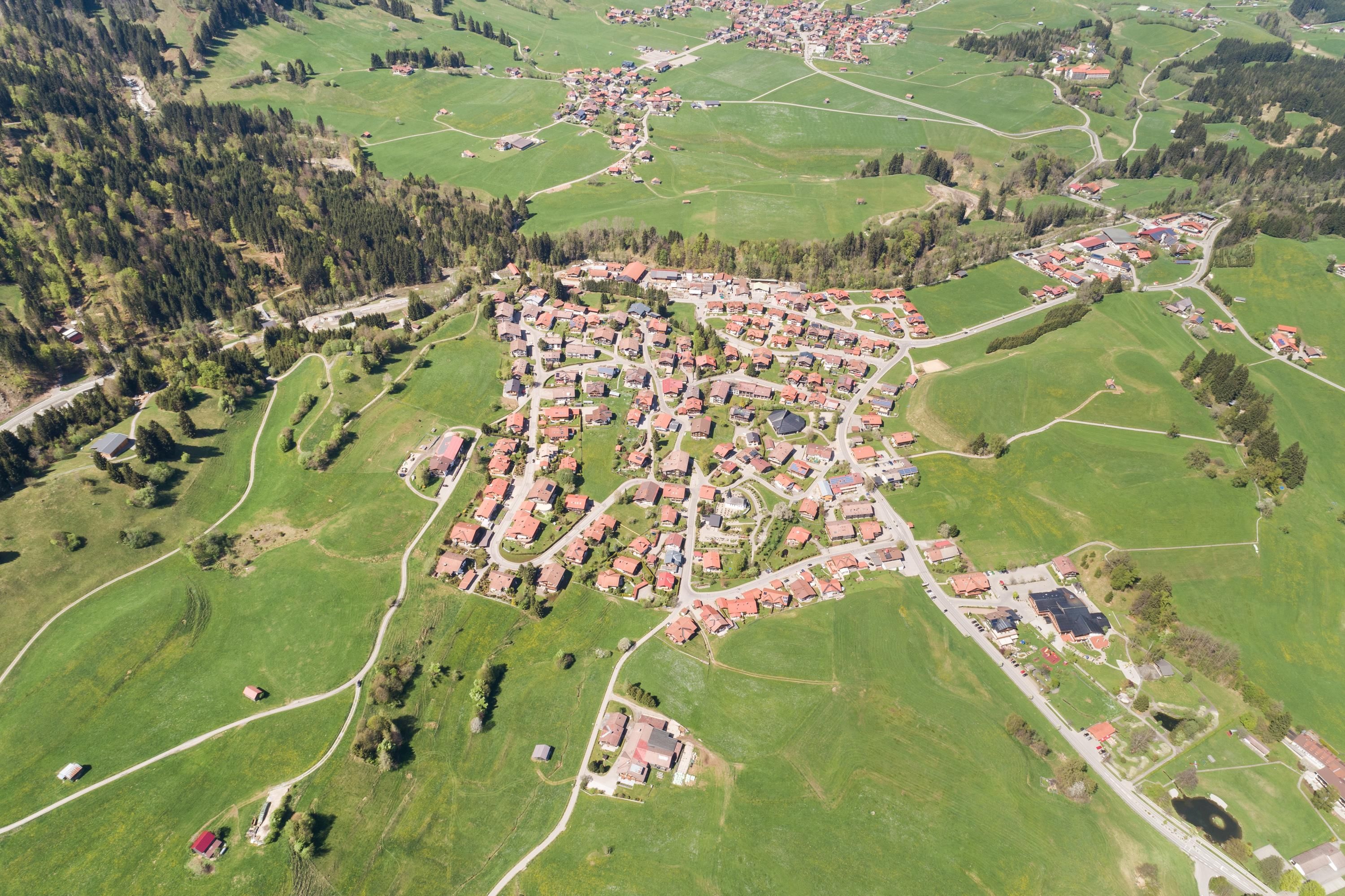 Vogelperspektive auf Parkplatz, umgeben von grünen Wiesen, Häusern, Straßen und bewaldeten Hängen.