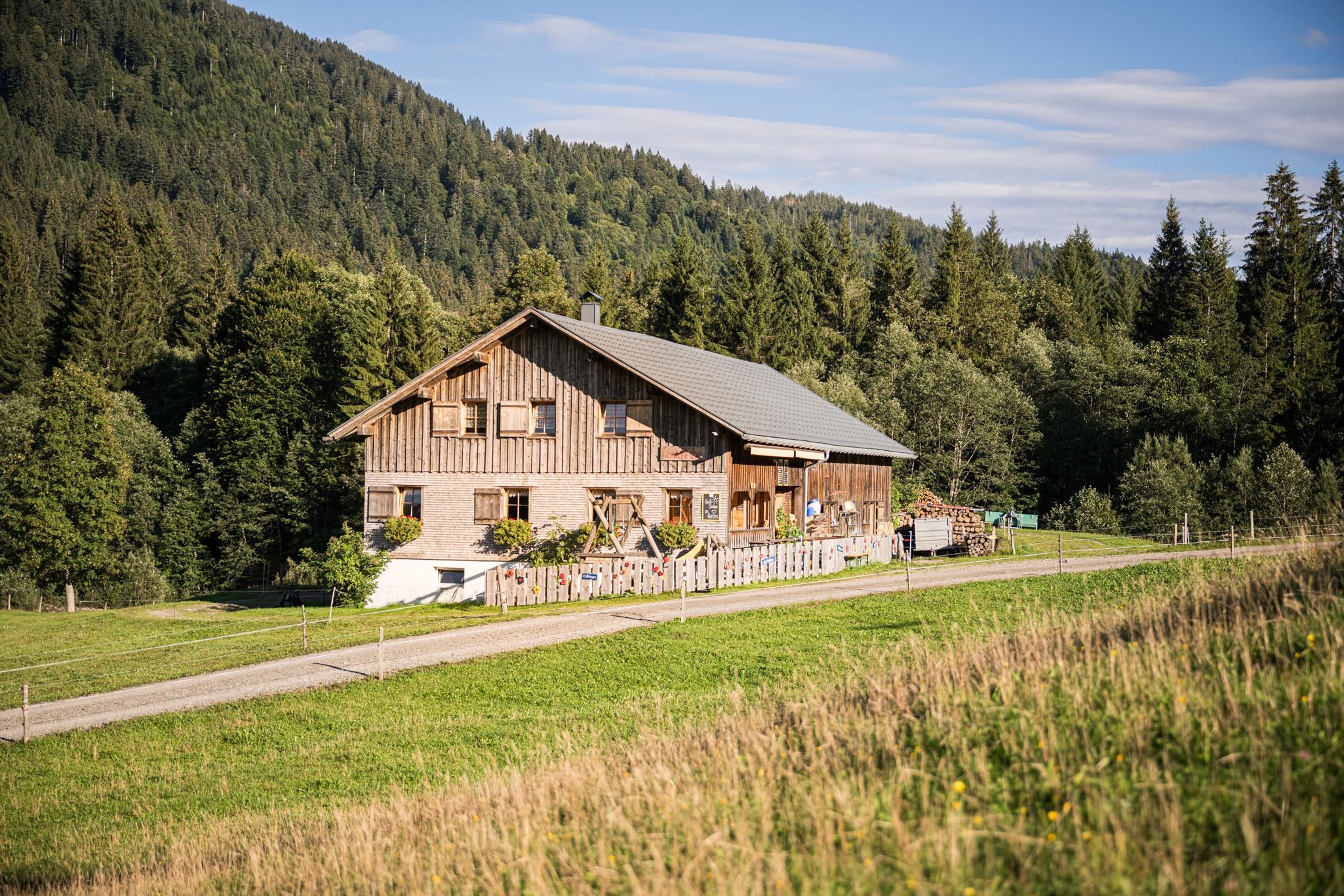 Ein rustikales Holzhaus in einer grünen Landschaft. Im Hintergrund sind Wälder und sanfte Hügel zu sehen.
