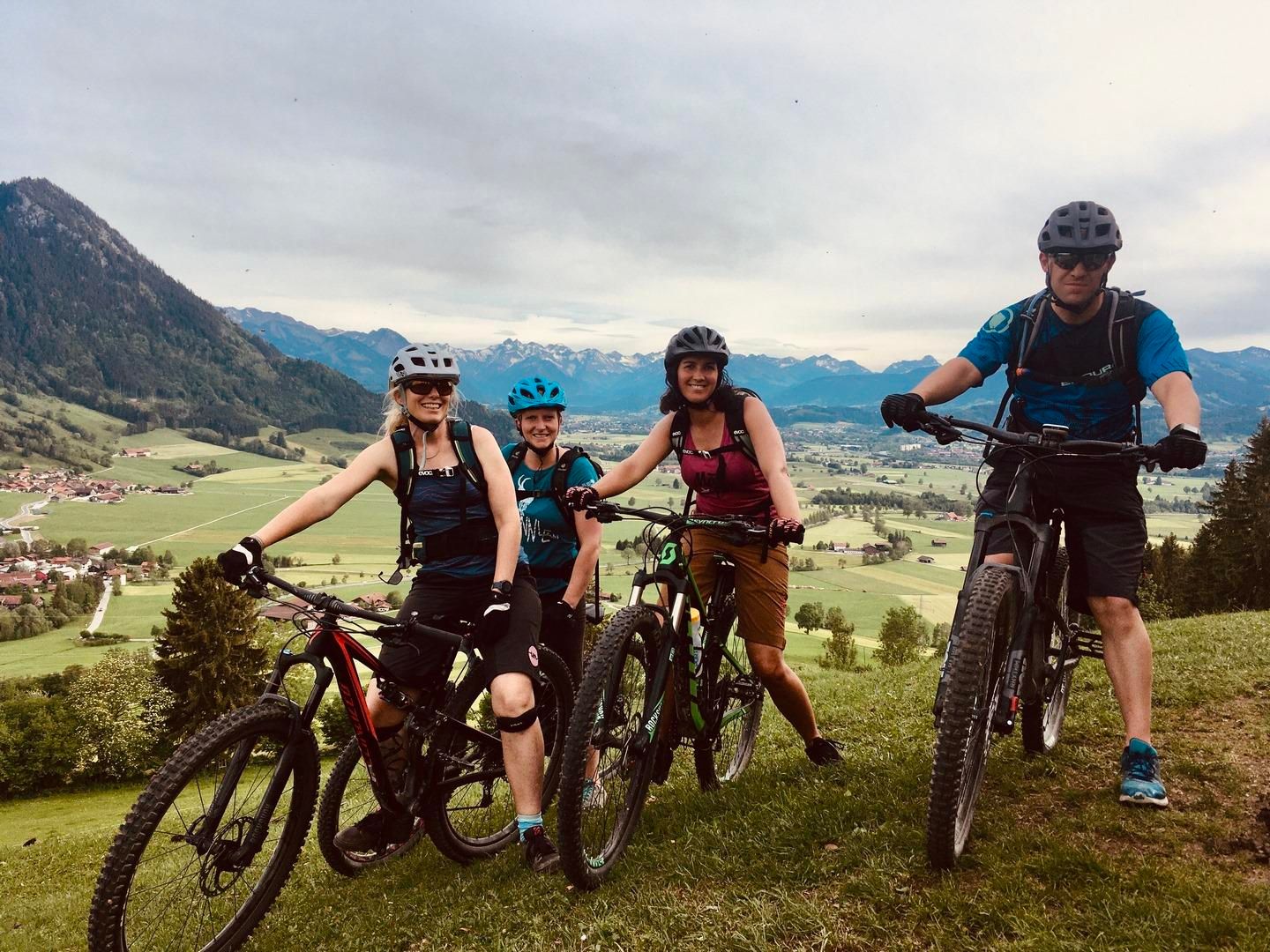 Vier Personen mit Mountainbikes und Helmen posieren auf einer grünen Wiese. Im Hintergrund eine weite Landschaft mit Bergen und einem bewölkten Himmel.