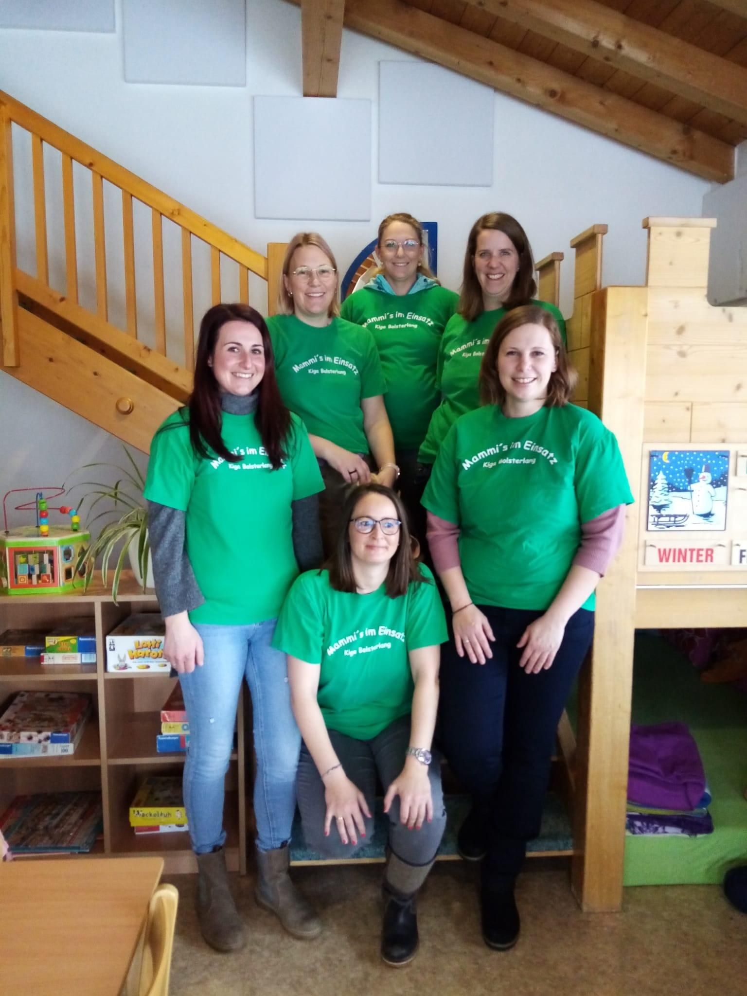 Sechs Frauen in grünen T-Shirts posieren auf einer Holztreppe. Sie lächeln in die Kamera. Bücherregale und Spielzeug sind im Hintergrund sichtbar.