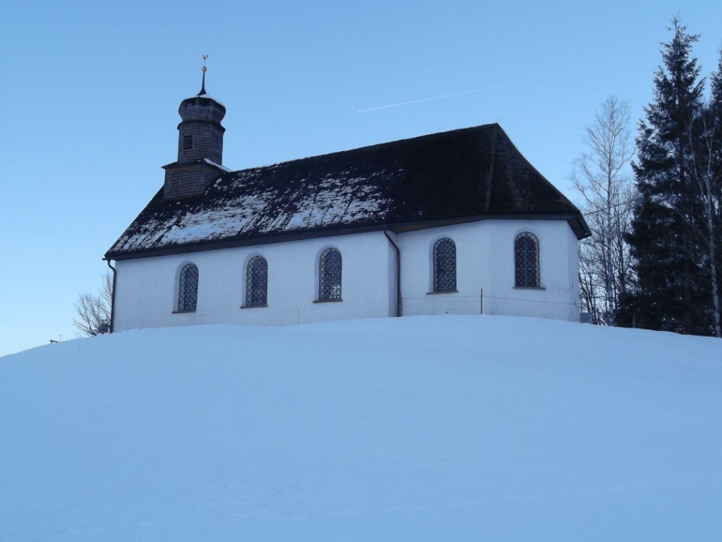 Kleine weiße Kapelle mit dunklem Dach auf einer schneebedeckten Anhöhe. Blauer Himmel im Hintergrund.