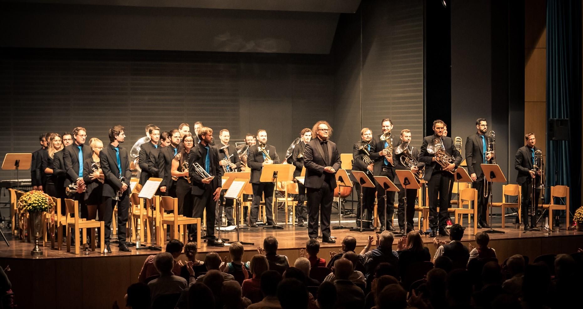 Brass Band A7 mit dem Dirigenten Thomas Wolf, alle mit schwarzer Kleidung, die Männer tragen türkisfarbene Krawatten.