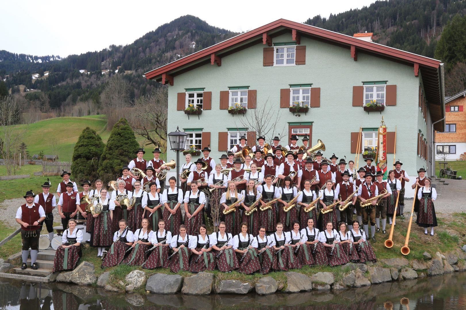 Eine große Gruppe in Trachten steht vor einem traditionellen Haus mit Teich.