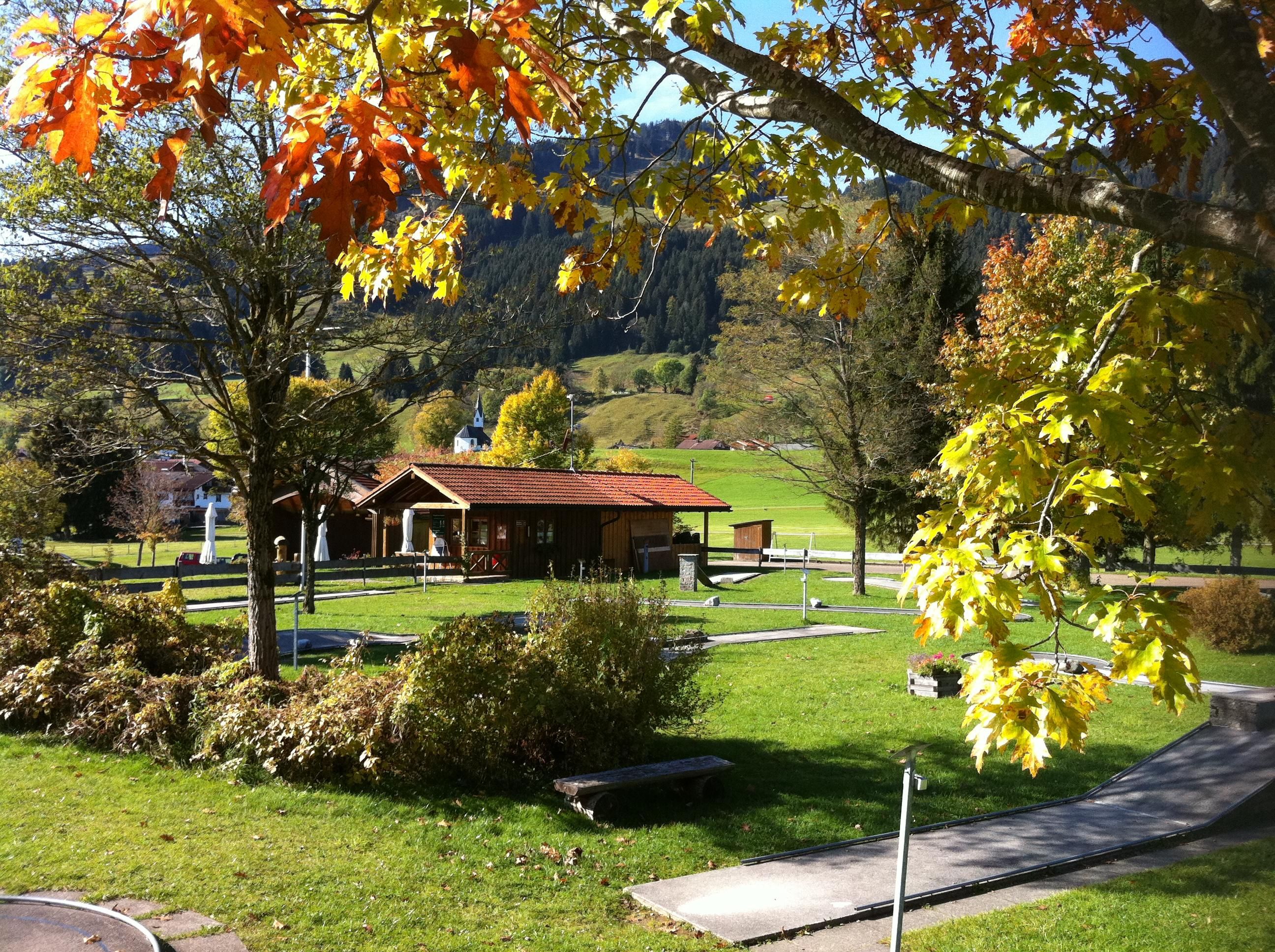 Herbstliche Ansicht von Schickie's Minigolfanlage mit bunten Blättern an den Bäumen, grünen Minigolfbahnen und einem hölzernen Häuschen.