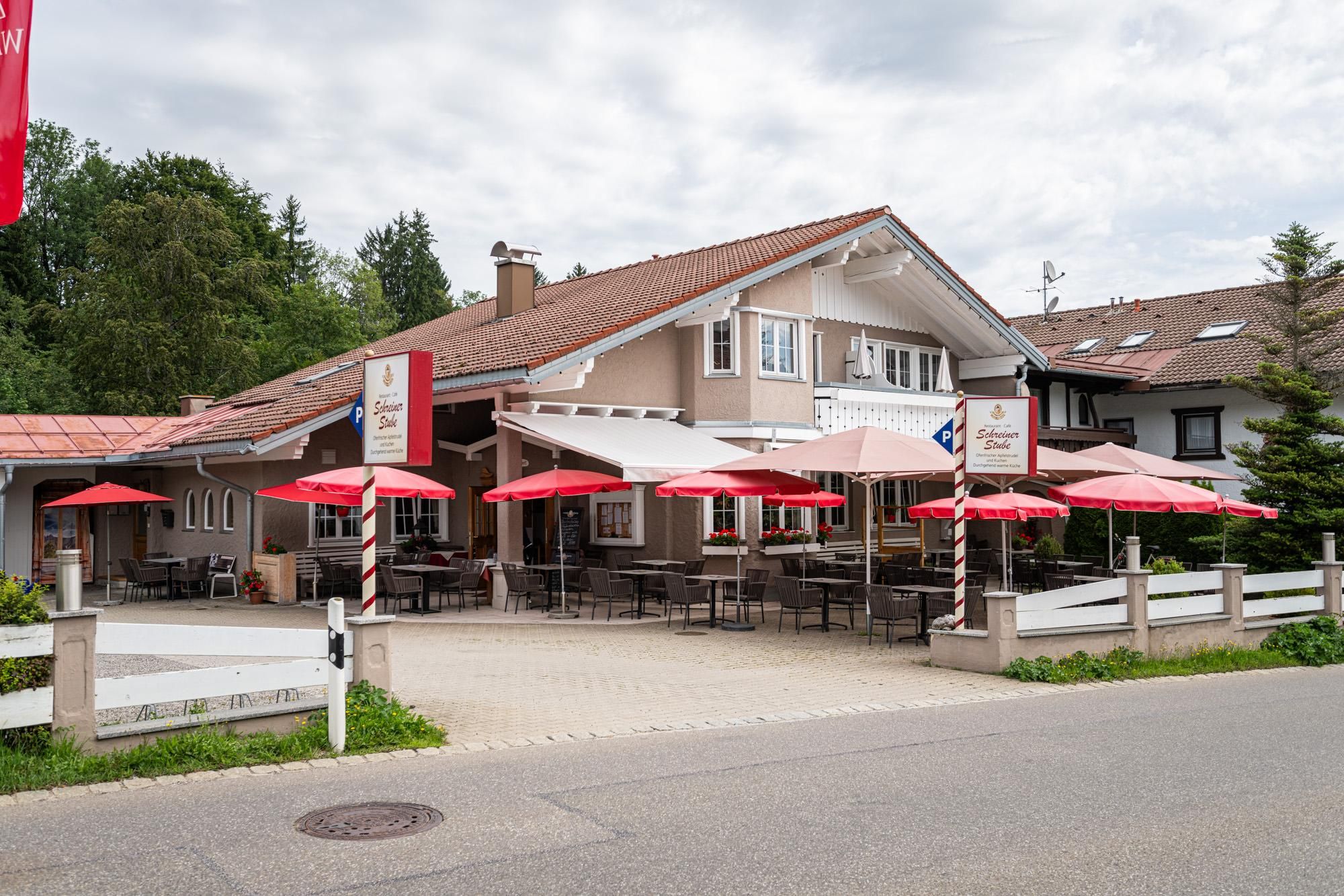 Beige Gebäude mit braunem Dach und roten Sonnenschirmen vor der Terrasse. Tische und Stühle im Freien. Schilder mit dem Namen des Restaurants.