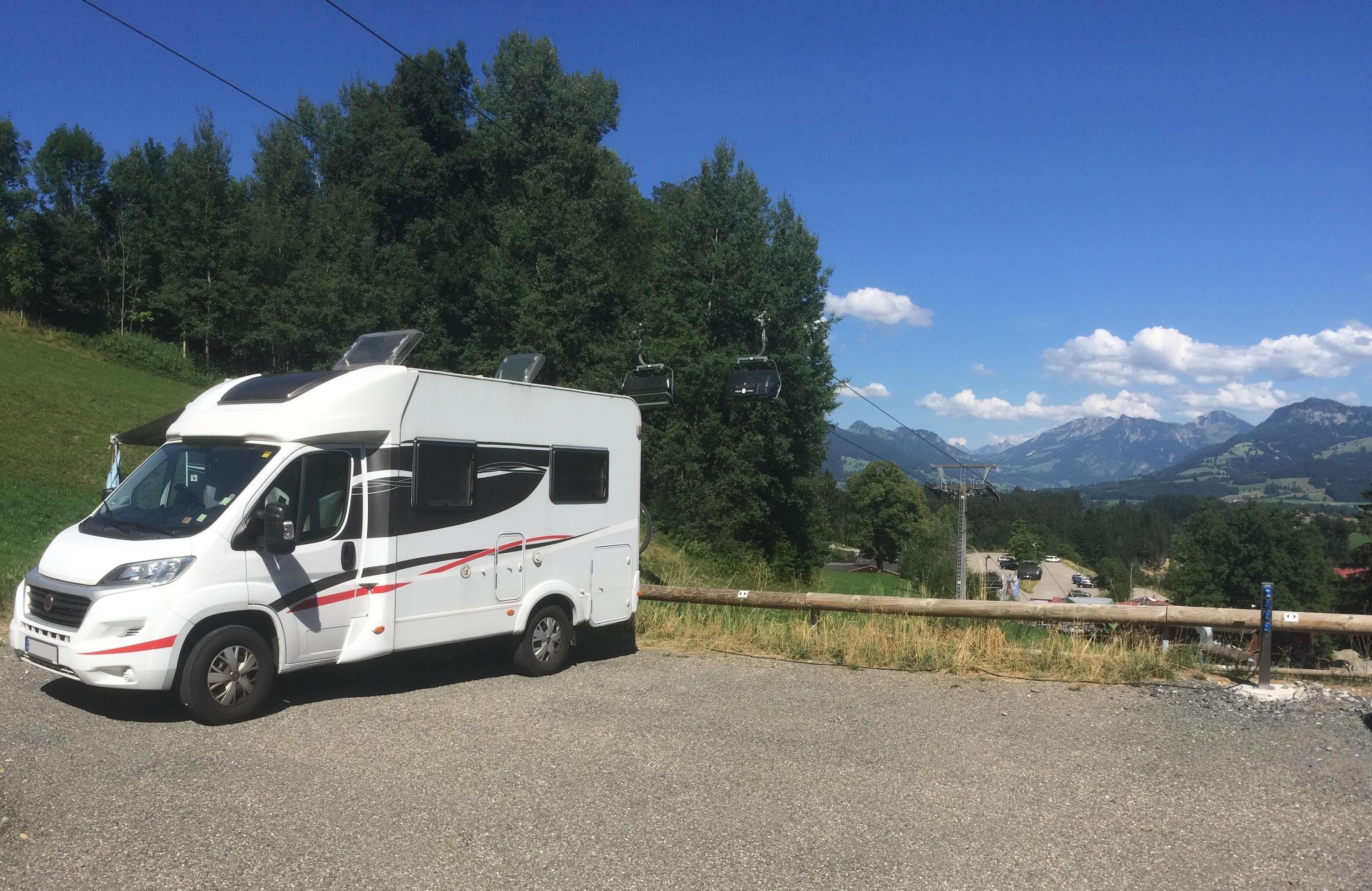 Weißes Reisemobil, möglicherweise ein Sunlight T68, parkt auf Schotterplatz. Im Hintergrund grüne Landschaft, Bergpanorama und eine Sesselbahn unter blauem Himmel mit Wolken.