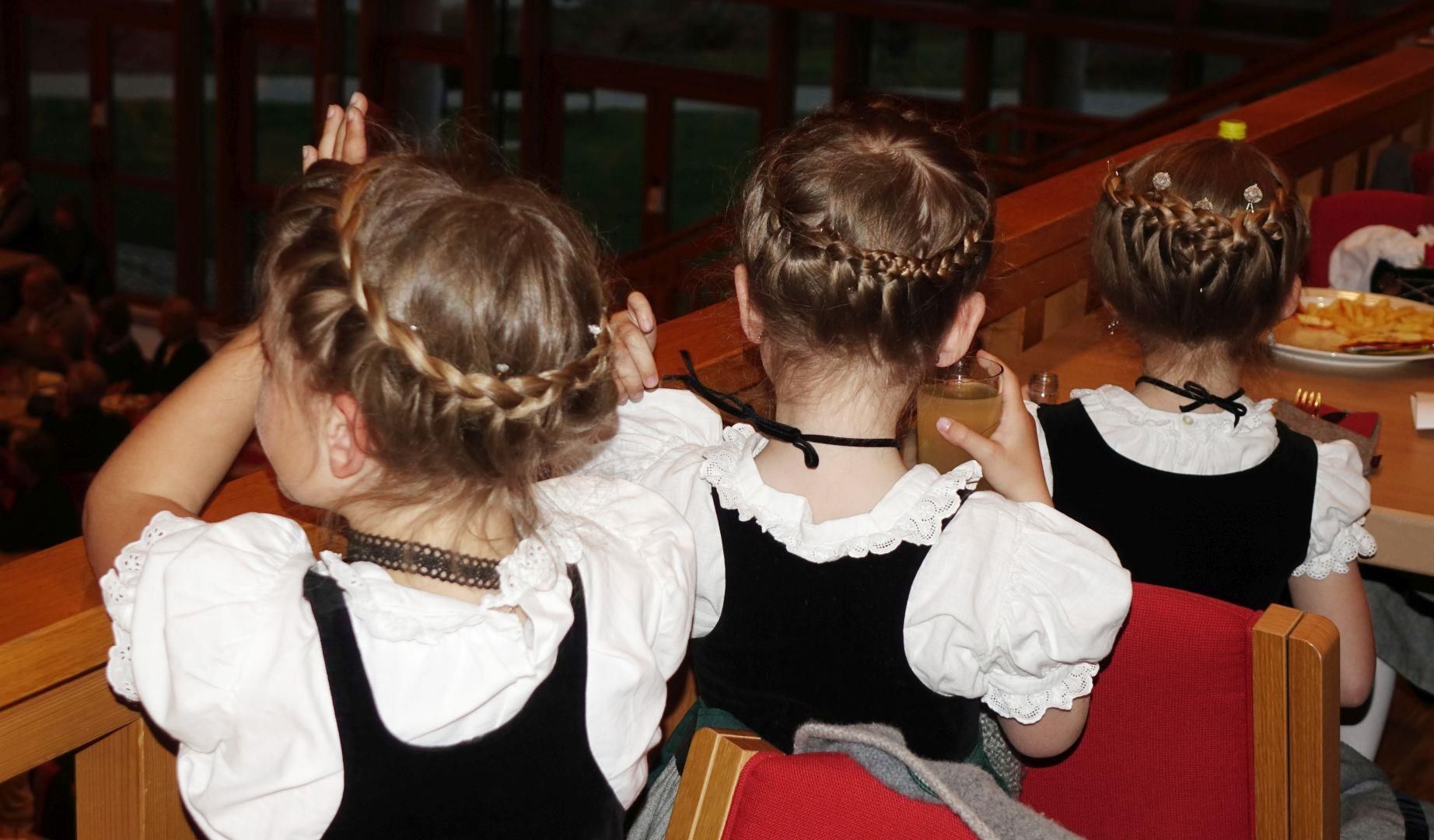 Drei Kinder, von hinten fotografiert, mit geflochtenen Haarkränzen, weißen Blusen und schwarzen Dirndloberteilen.