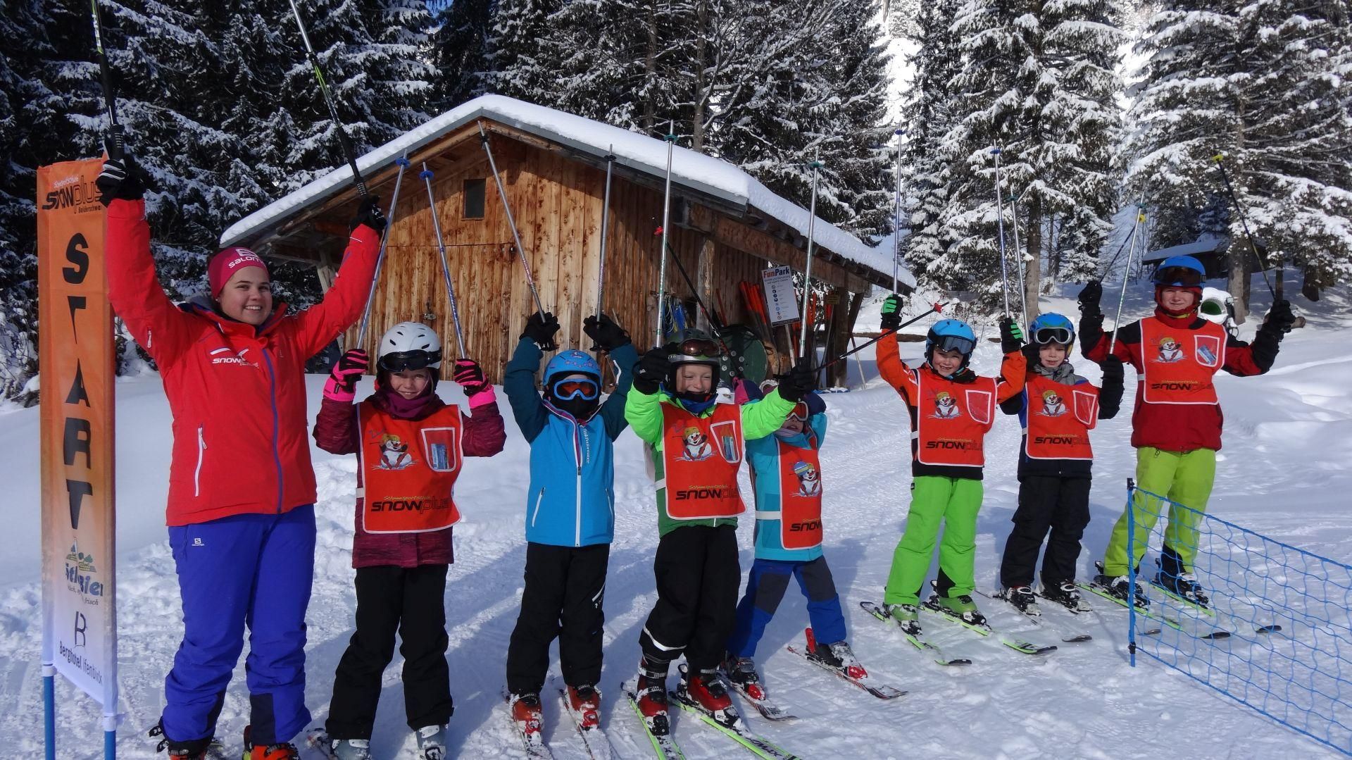 Skilehrerin und sechs Kinder mit Skiern und Helmen jubeln vor einer Holzhütte im Schnee, "START" Schild sichtbar.
