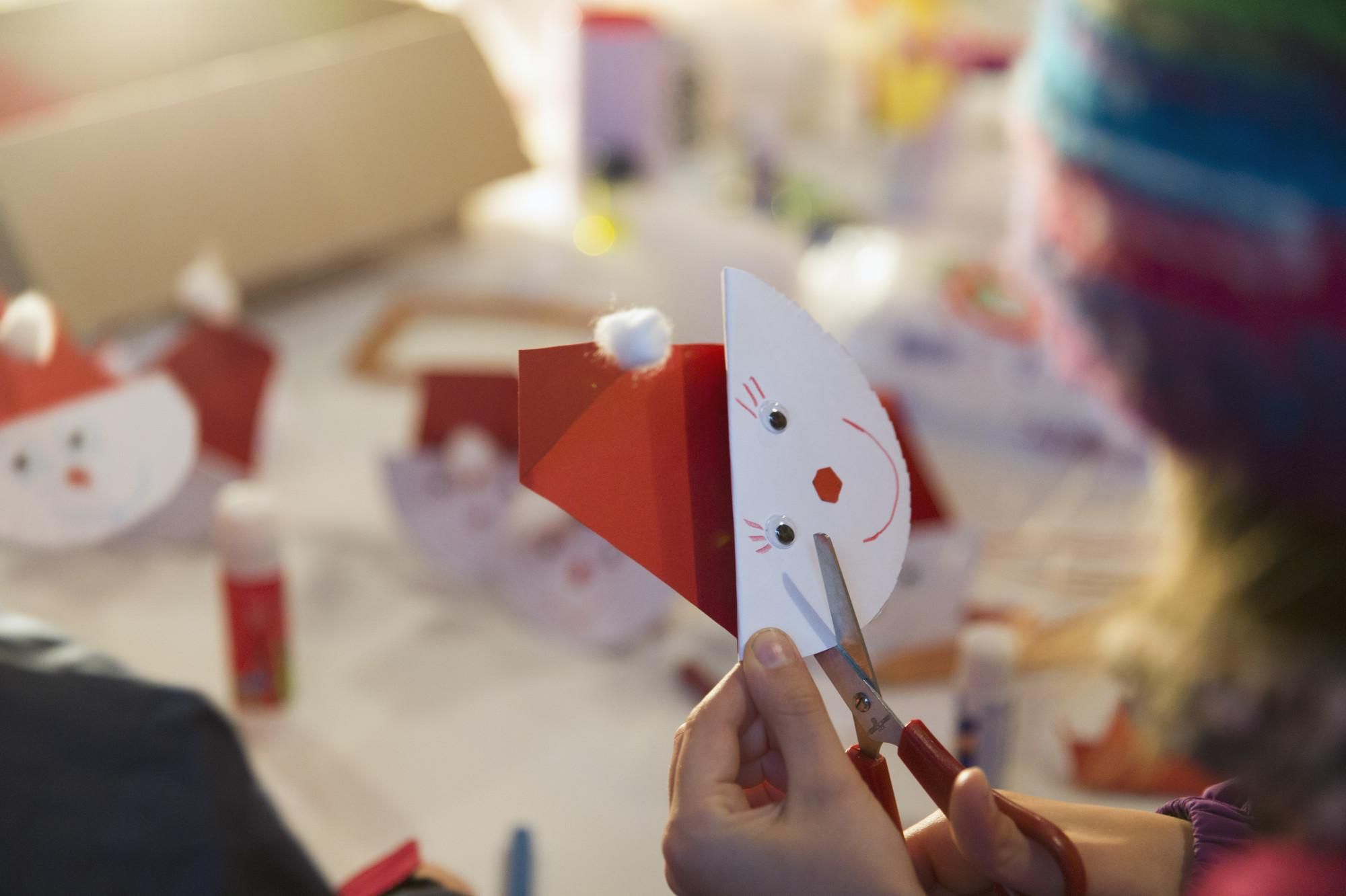 Ein Kind schneidet mit einer Schere eine Weihnachtskarte in Form eines Schneemanns. Im Hintergrund sind weitere Bastelmaterialien zu sehen.