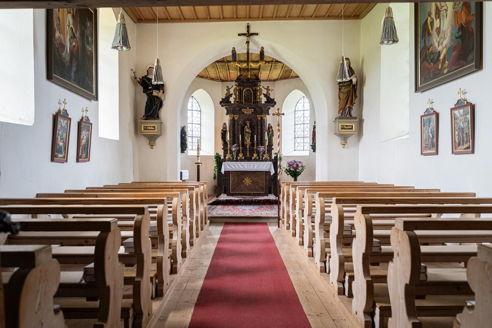Innenansicht einer schlichten Kapelle mit Holzbänken und einem verzierten Altar unter einem Holzkreuz. Ein roter Teppich liegt auf dem hellen Holzboden.