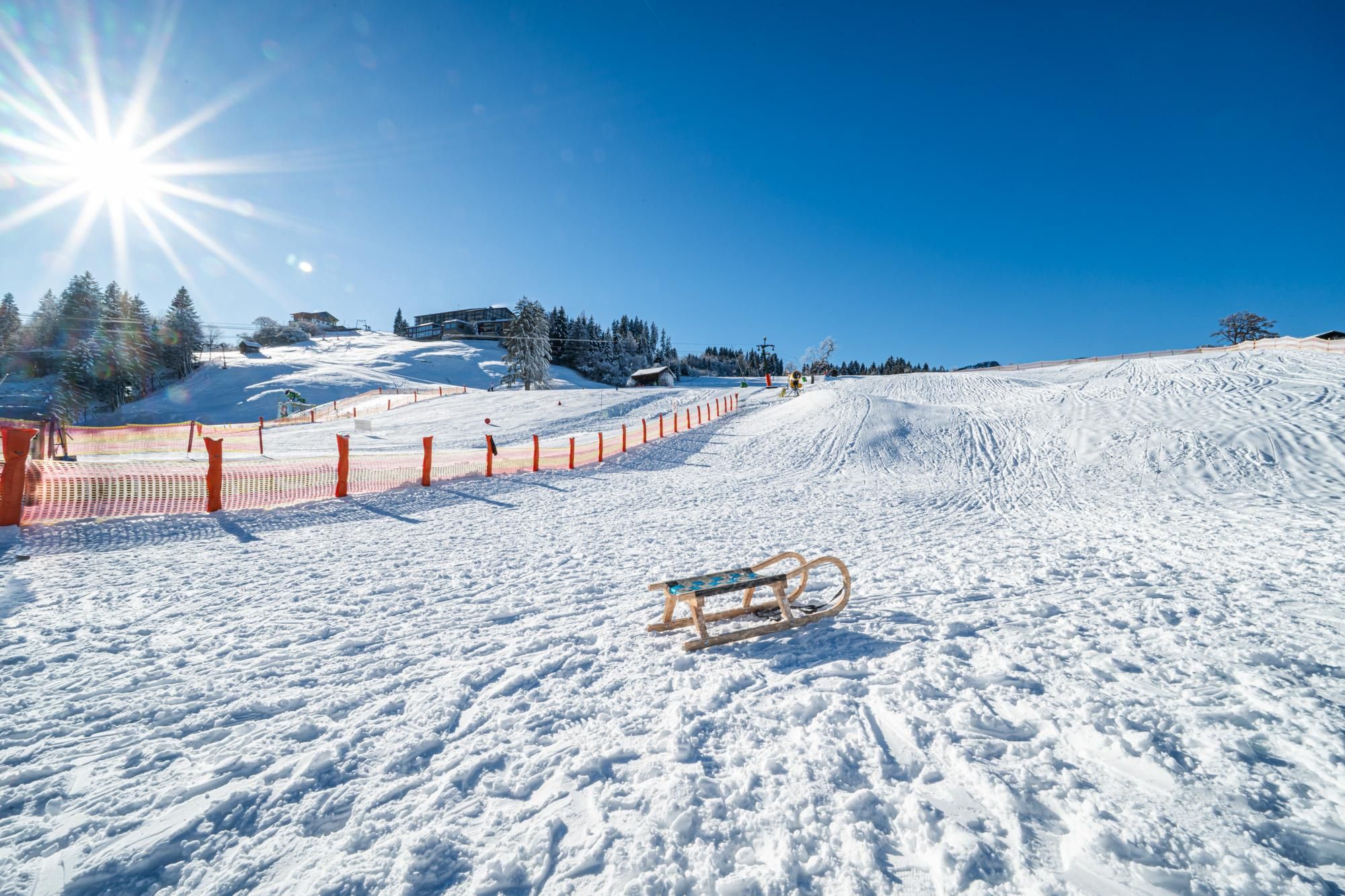 Sonnige Rodelpiste am Stinesser in Fischen mit klassischem Holzschlitten im Schnee unter strahlend blauem Himmel.