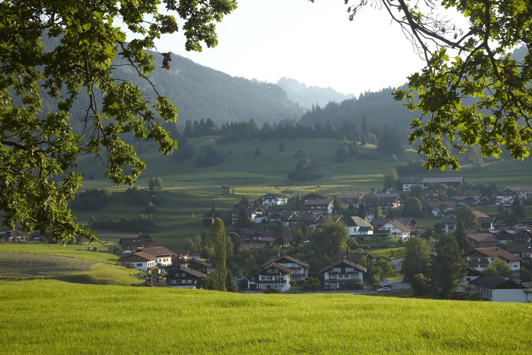Obermaiselstein: Blick durch grüne Äste auf Dorf mit Häusern in grüner Landschaft, Berge im Hintergrund, heller Himmel.