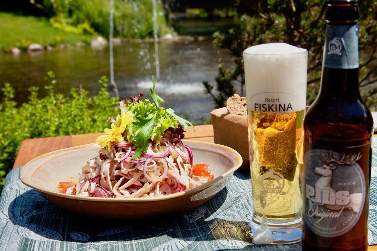 Auf einem Terrassentisch steht ein Teller mit Wurstsalat, dazu ein Pils, im Hintergrund der Grundbach mit Springbrunnen.
