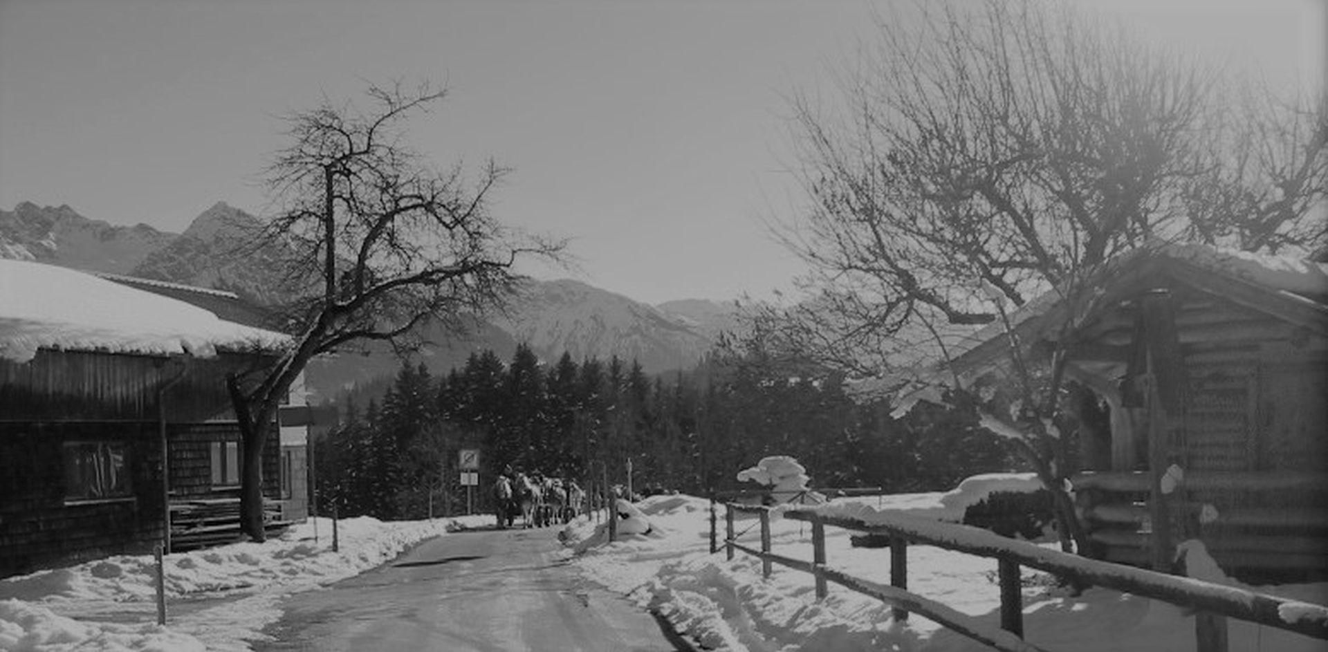 Schwarz-Weiß-Aufnahme einer verschneiten Winterlandschaft mit Häusern, Bäumen und Bergen im Hintergrund. Eine Gruppe von Personen geht auf der geräumten Straße.