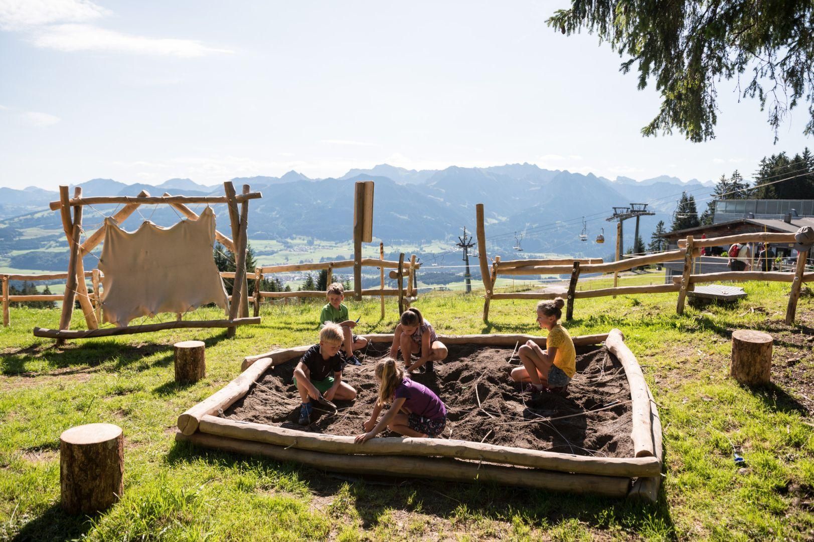 Sechs Kinder spielen in einem Sandkasten aus Baumstämmen.