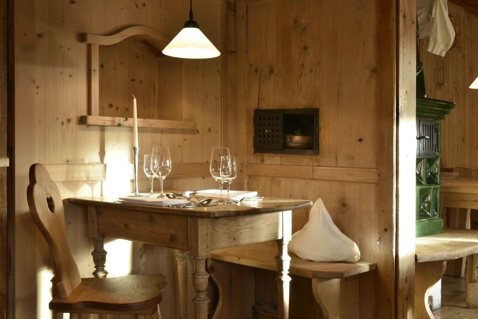 Gemütliche Ecke mit Holztisch und Stühlen. Gedeckt mit Weingläsern, Besteck und Kerze. Holzvertäfelte Wände, eine Hängelampe und grüner Kachelofen im Hintergrund.