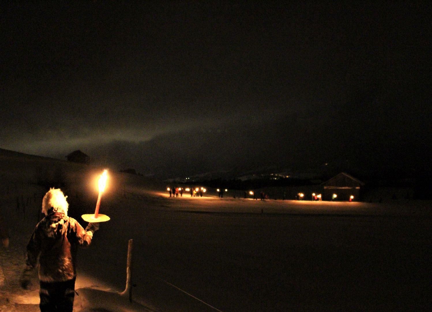 Dunkle Nacht: Ein Kind hält eine Fackel, im Hintergrund weitere Lichter und Häuser im Schnee.