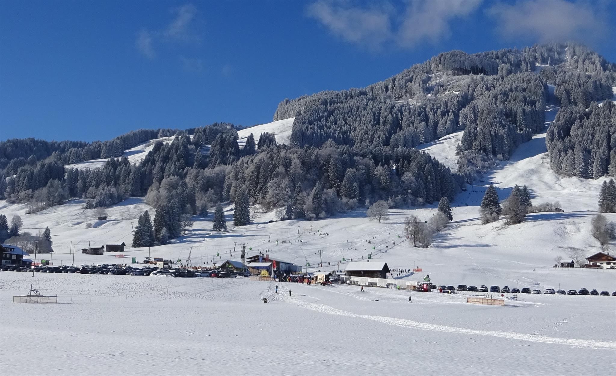 Verschneite Winterlandschaft am Basislager Bolsterlang mit Skifahrern, Liften und Parkplätzen unter einem klaren blauen Himmel mit einzelnen Wolken.