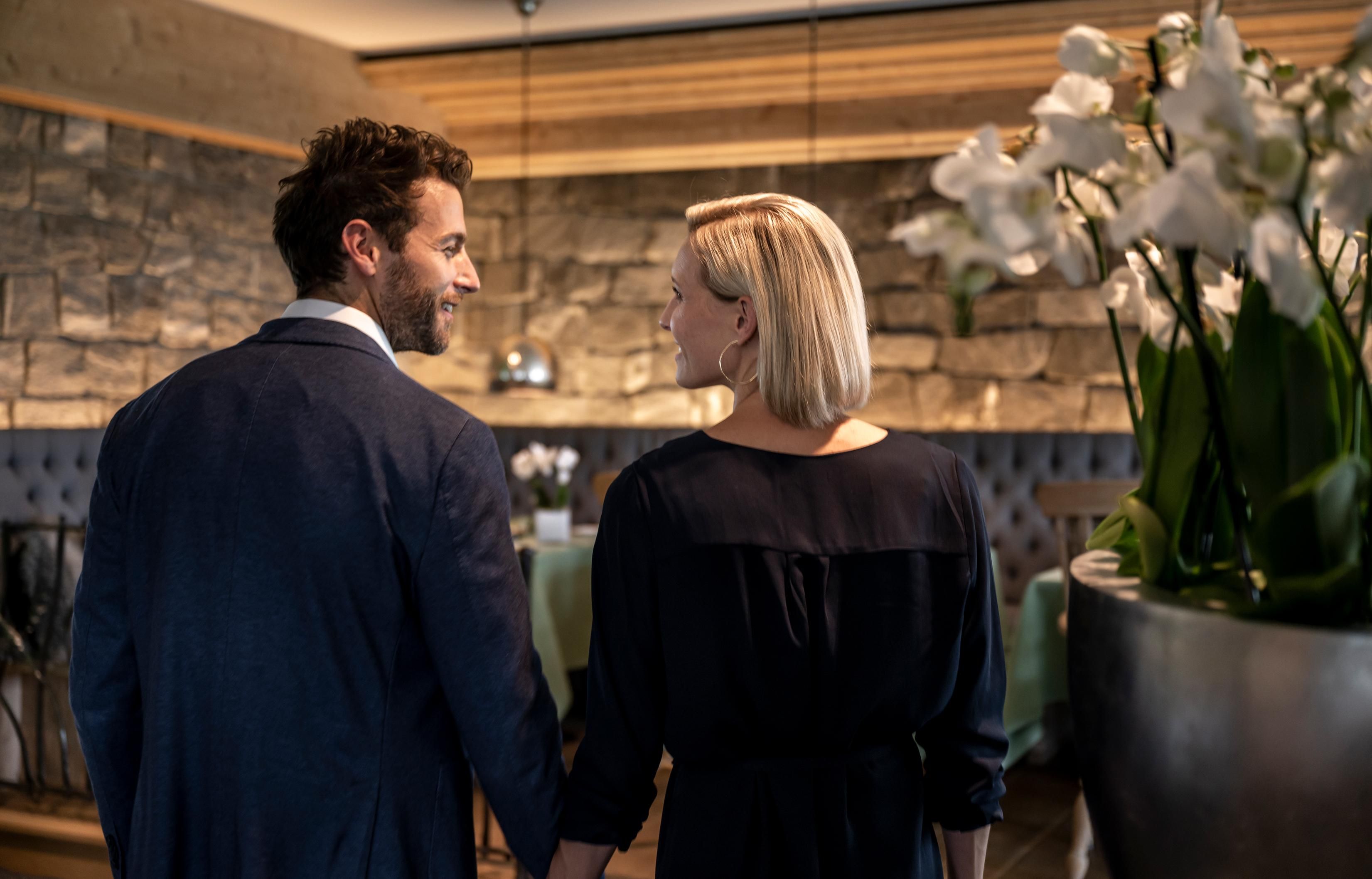 Paar lächelt sich im Restaurant an, sie trägt dunkles Kleid, er Anzug, Orchideen im Vordergrund.