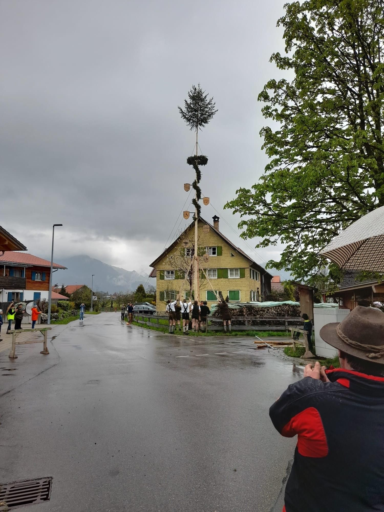 Dorfszene mit Maibaum und Menschen in Tracht vor Haus, grauer Himmel und Regen.