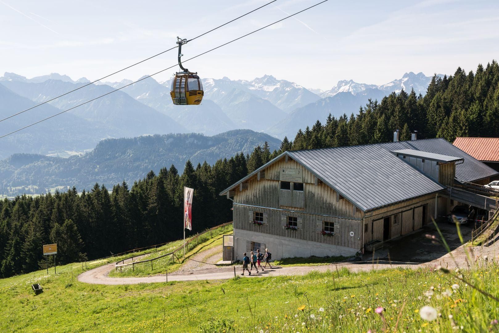 Hölzerne Sennalpe mit graublauem Dach auf grüner Bergwiese, Seilbahnkabine schwebt darüber, Wanderer unterwegs, Alpenpanorama im Hintergrund.