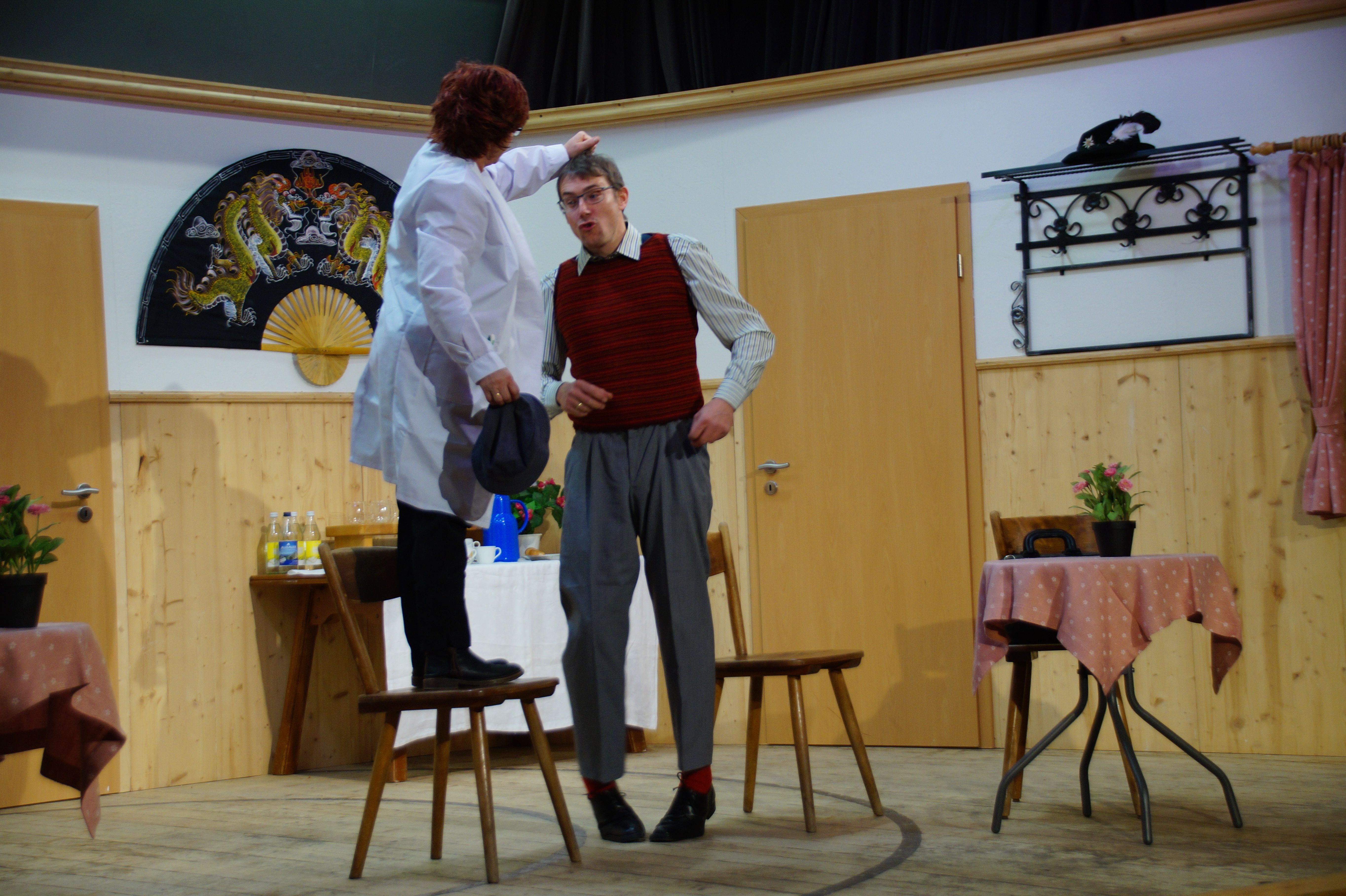 Eine Frau im weißen Arztkittel steht auf einem Stuhl und zieht einem Mann an den Haaren.