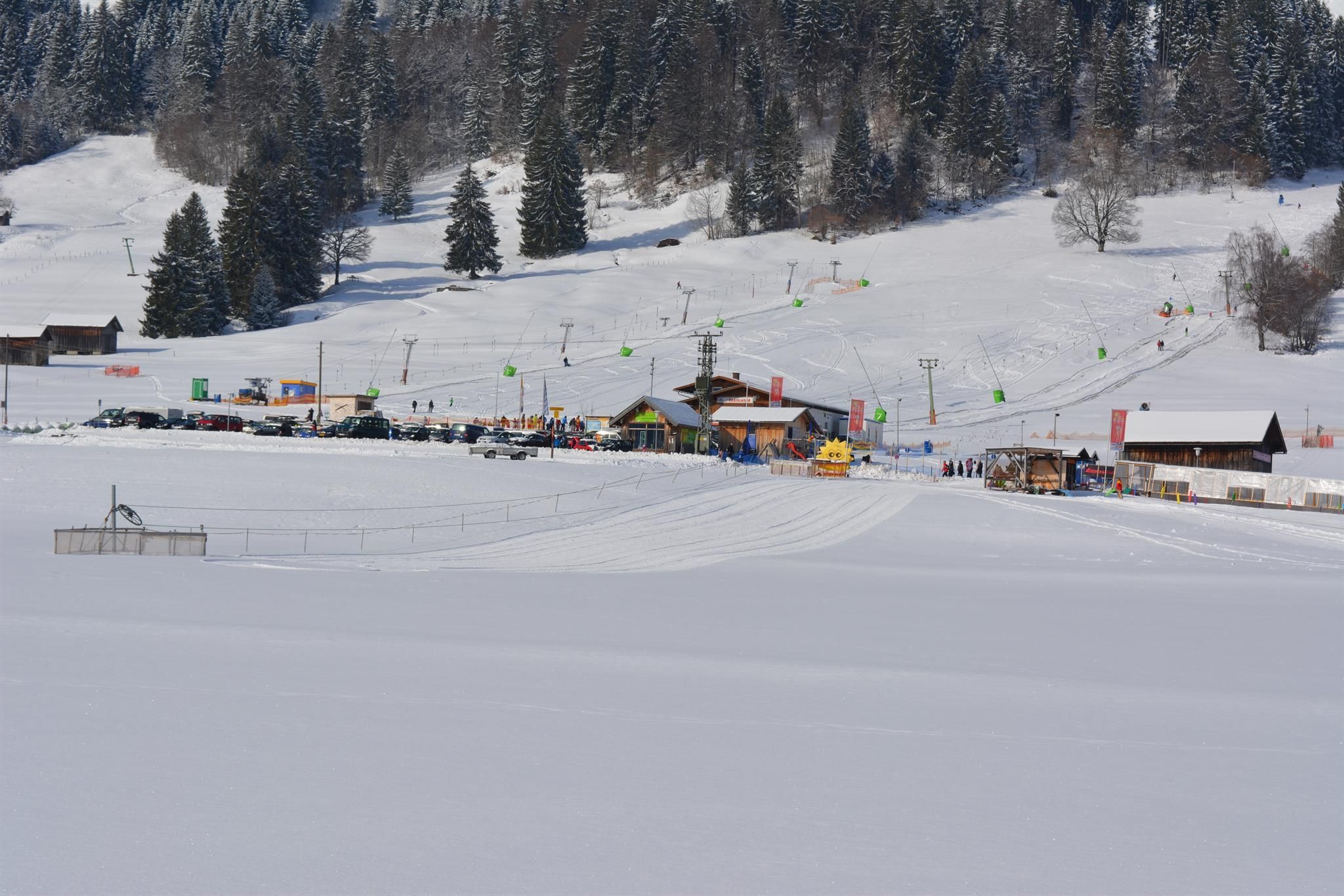 Winterlandschaft am Basislager Bolsterlang mit Skiliften, Skifahrern, Gebäuden und Parkplätzen unter einem leicht bewölkten Himmel.