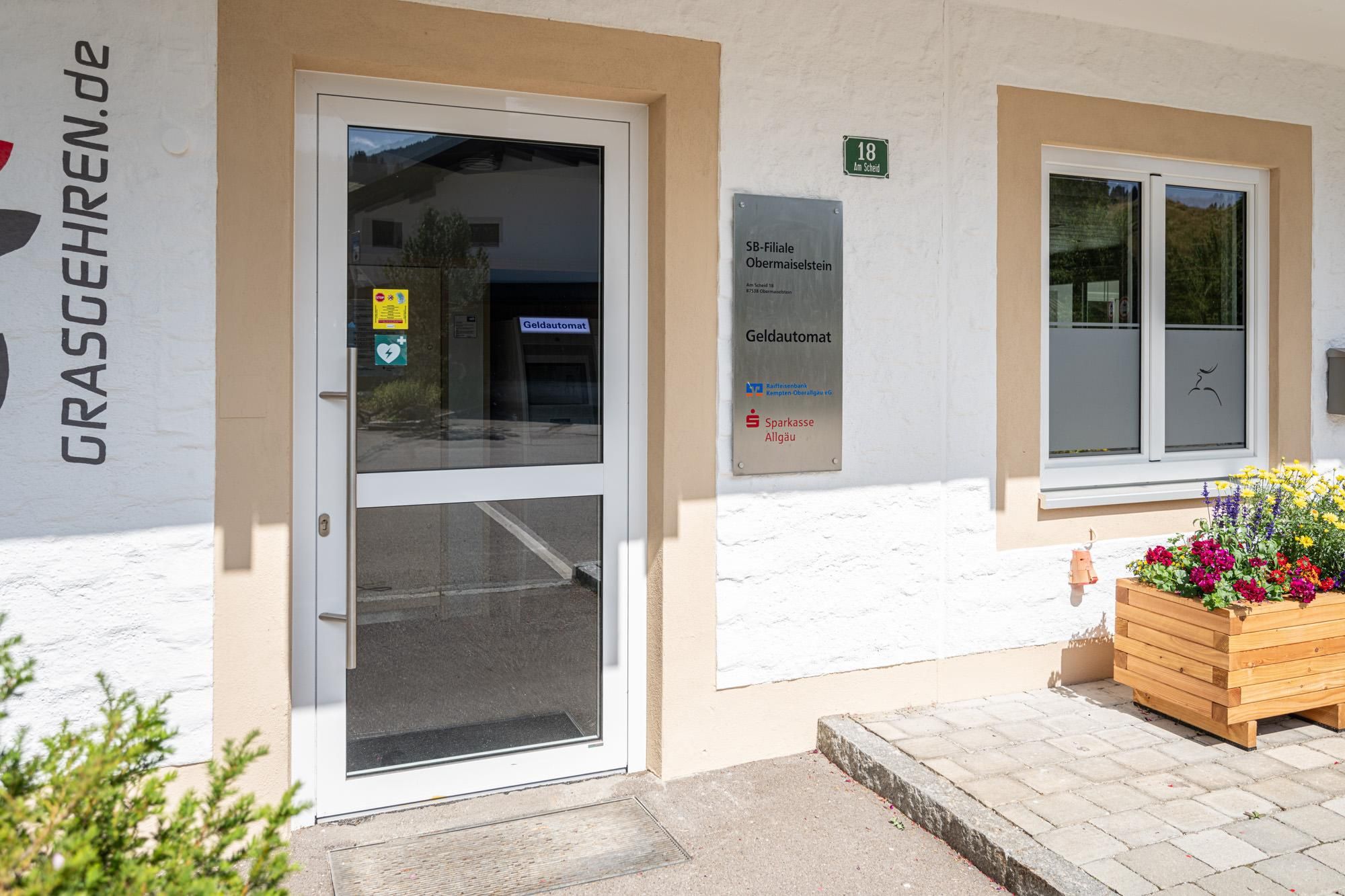 Eingangsbereich eines Geldautomaten in Obermaiselstein. Glastür mit Aufklebern. Schild mit Aufschrift "Geldautomat".