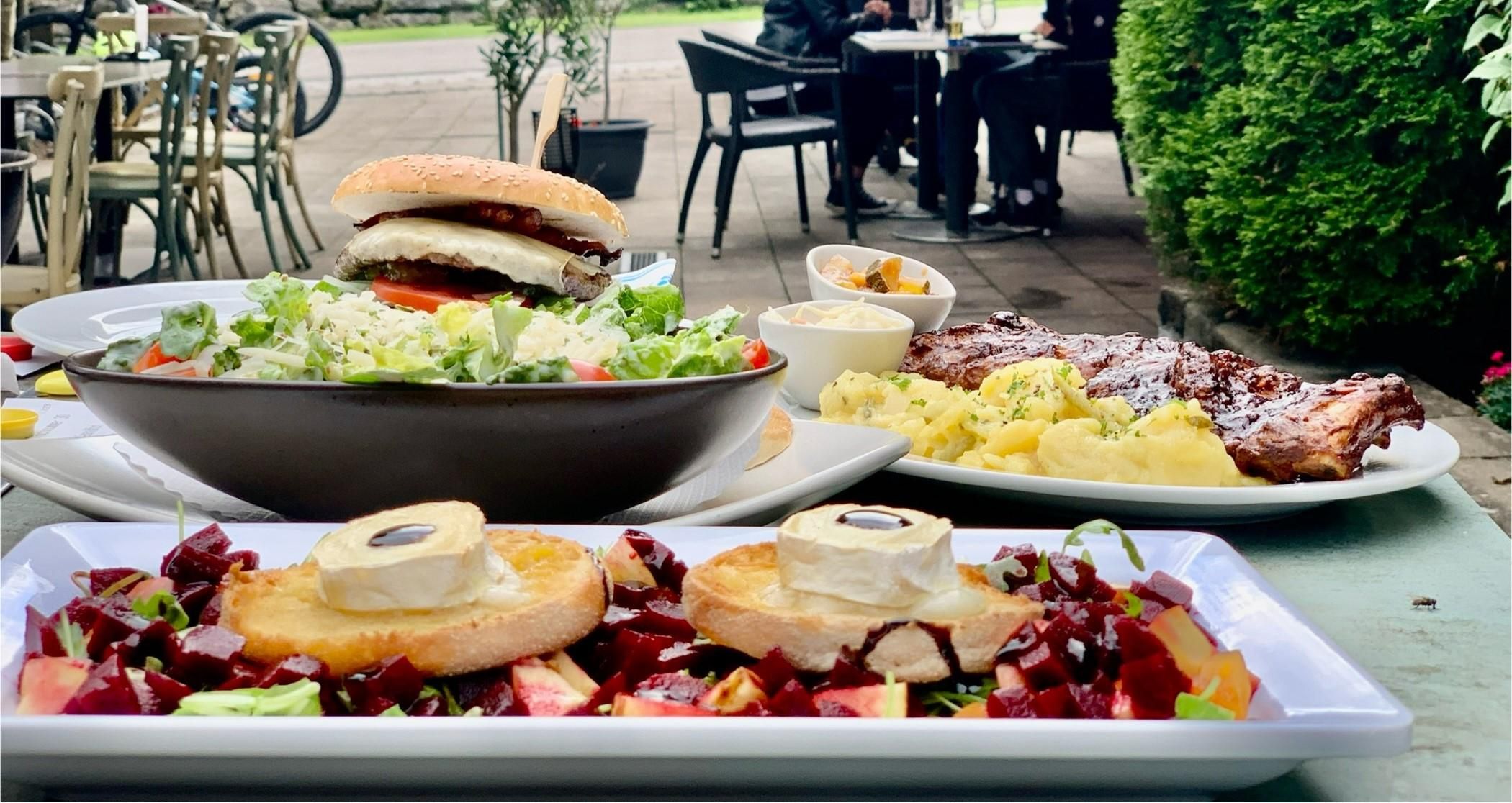 Nahaufnahme von verschiedenen Gerichten auf einer Terrasse. Ein Burger, Salate, Fleisch und Beilagen sind appetitlich angerichtet.