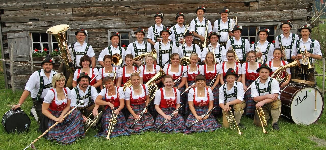 Weiße Kapelle mit spitzem, dunklem Turm auf einem grünen Hügel. Rechts ein großer Baum. Im Hintergrund sind Häuser.