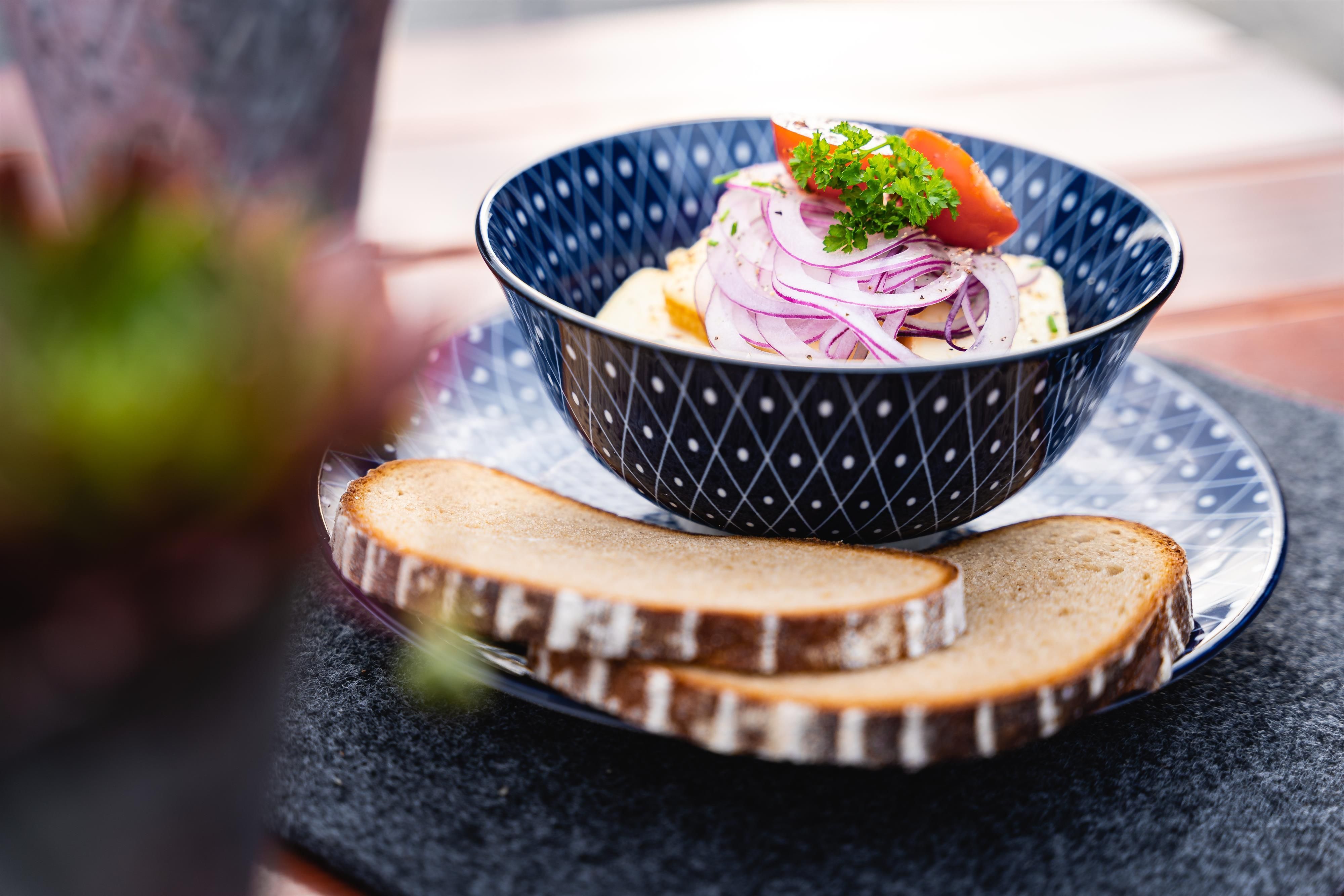 Obatzda in Schale: Bayrische Käsecreme mit Zwiebeln, Paprika, Petersilie, zwei Brotscheiben auf Teller, dunkler Untersetzer.
